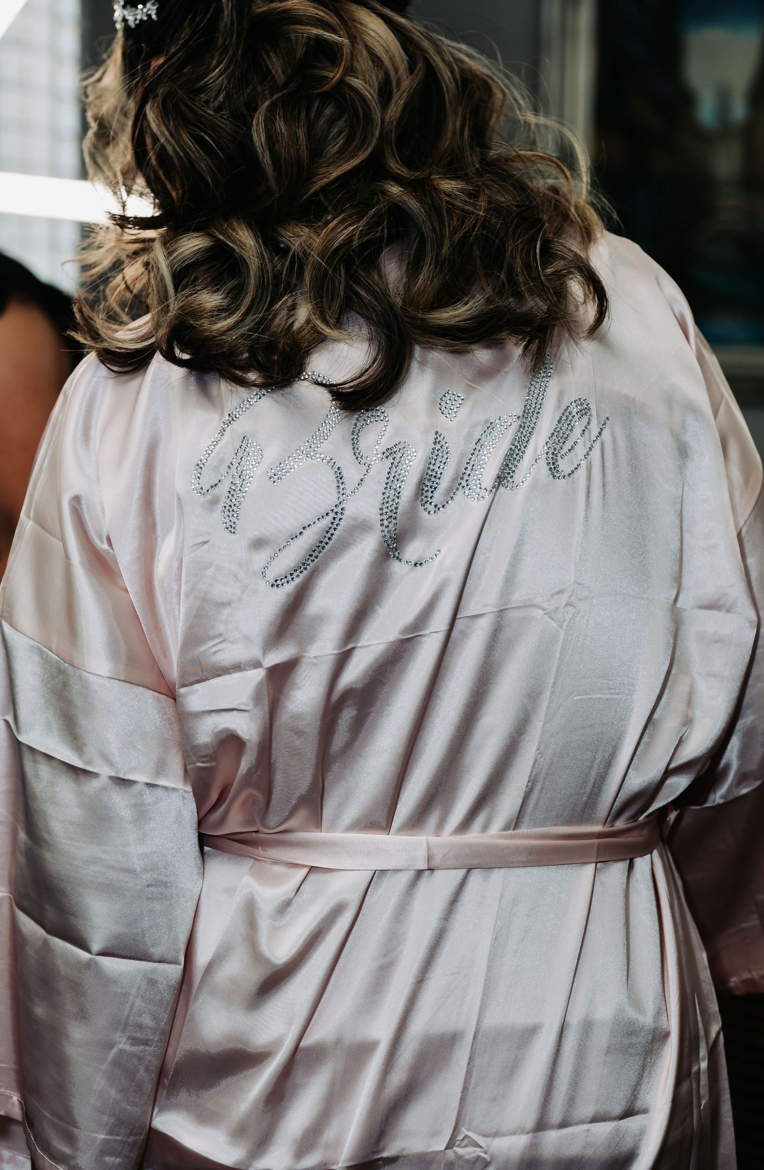 Bride wearing a pink satin robe with silver rhinestone script on the back, curly brunette hair, wedding preparation.