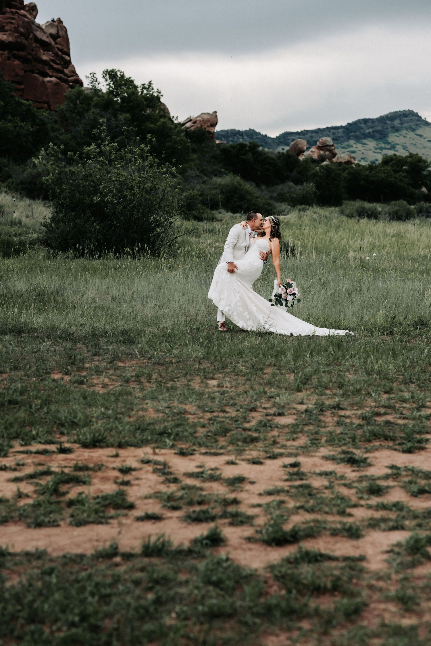 Bride and groom share a romantic dip kiss in a lush green meadow with red rock formations and mountains in the background.