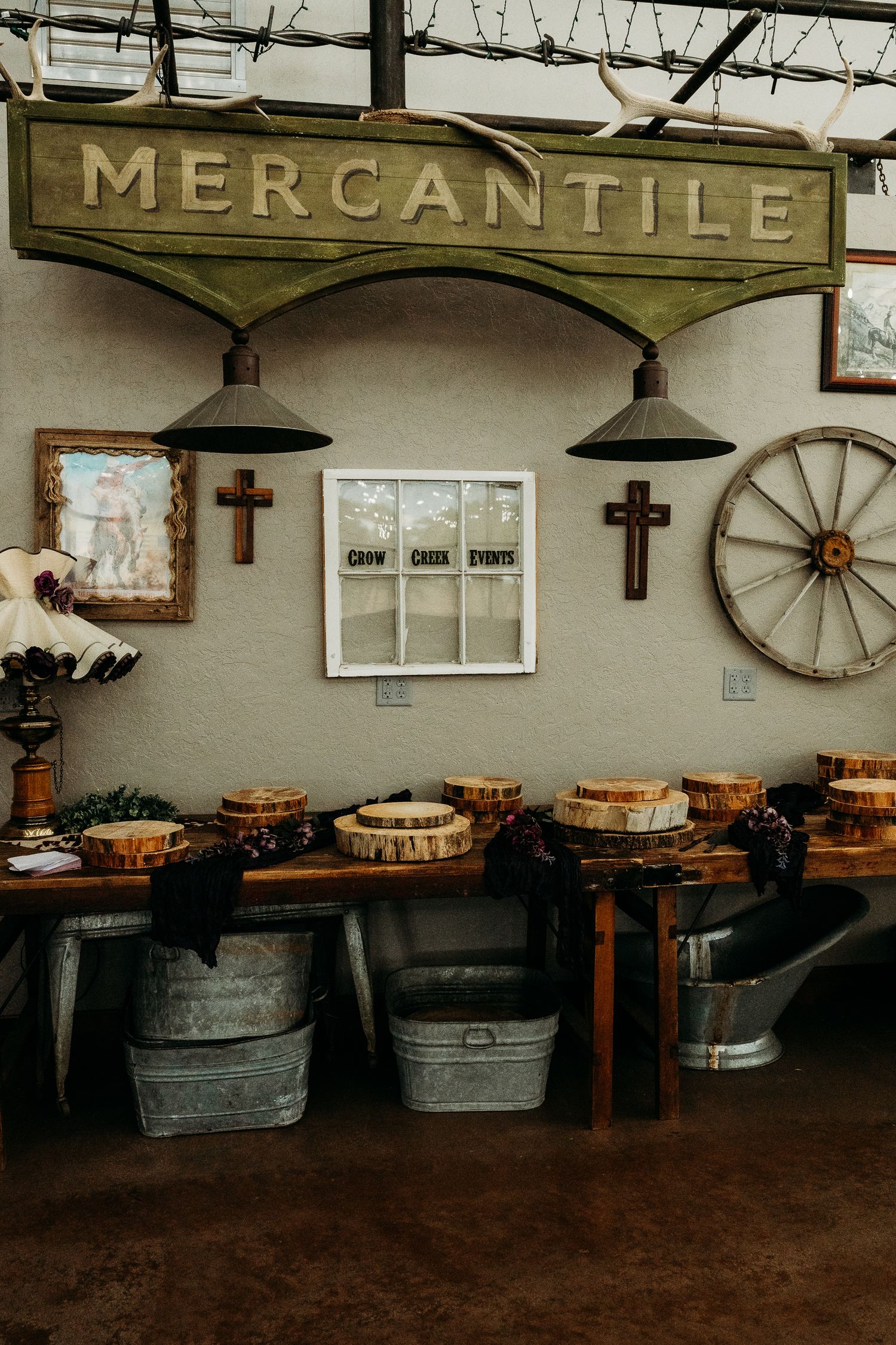 Rustic mercantile display with wooden sign, galvanized buckets, cross decor, and wagon wheel in a vintage-style event space.