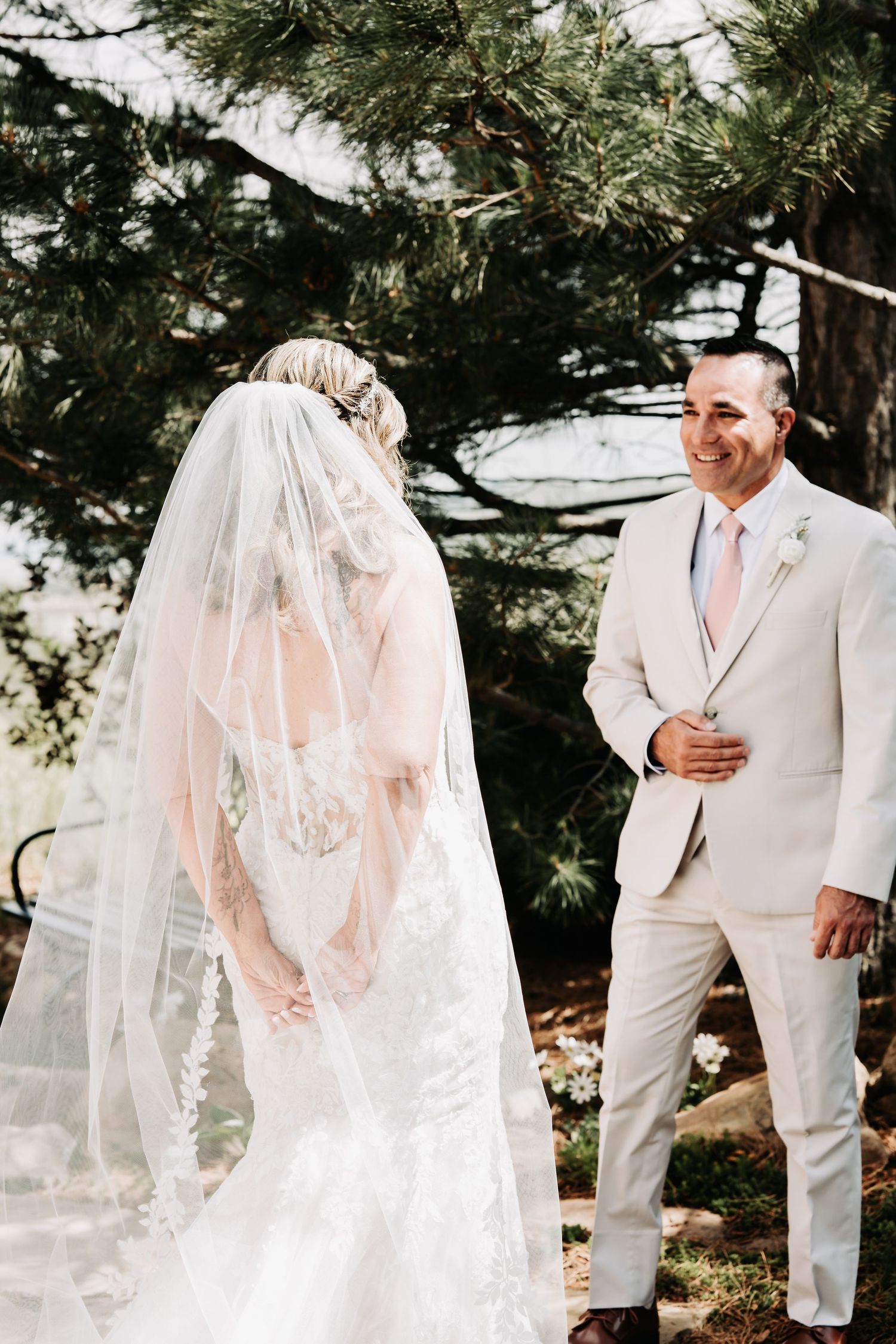 Groom in beige suit smiling at bride in lace wedding dress and veil during outdoor wedding ceremony under pine trees.