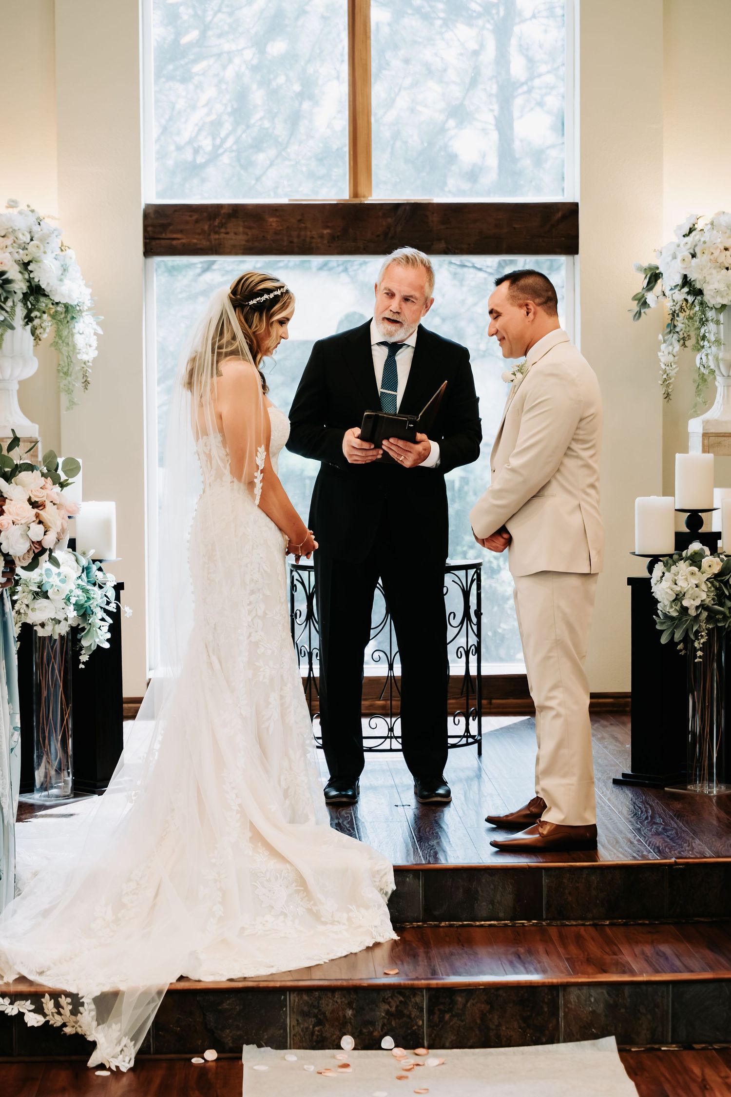 Bride and groom exchange vows with officiant at elegant indoor wedding ceremony with white floral arrangements.