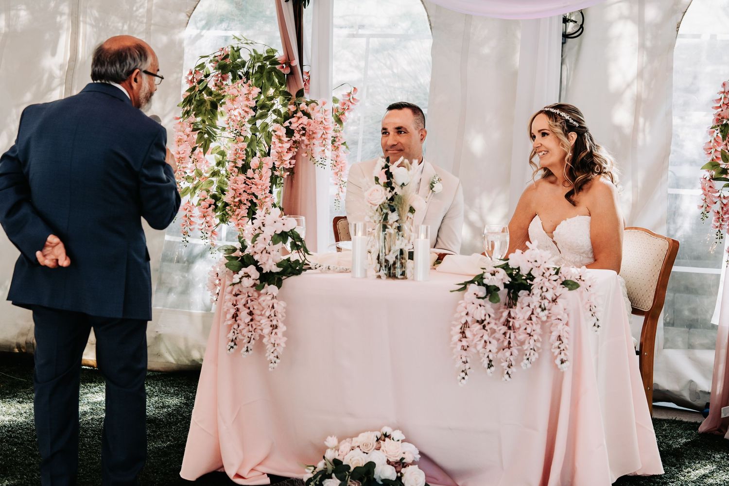 Bride and groom seated at sweetheart table with pink floral arrangements during wedding reception ceremony.