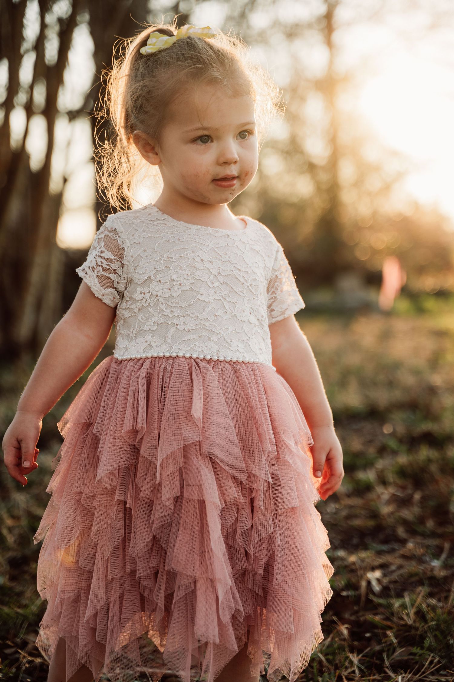 A toddler in a white lace and dusty pink tulle dress twirls in a sunlit forest setting during golden hour.