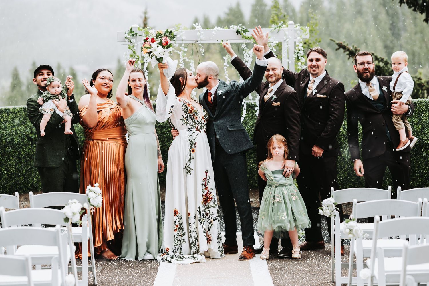Joyful wedding party celebrating outdoors under floral arch, bride and groom kissing, bridesmaids in sage green dresses.