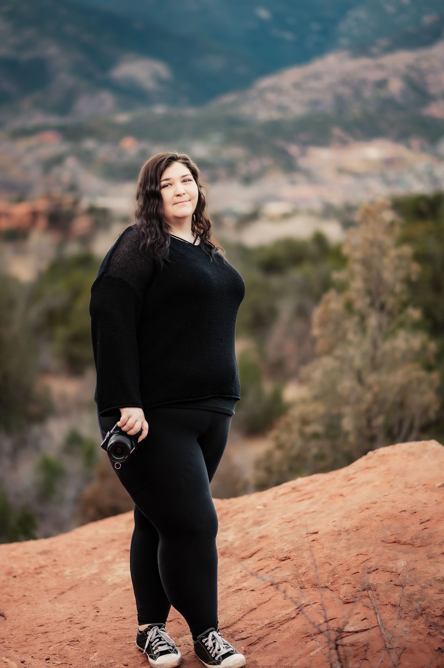 Woman in black outfit holding camera stands on red rocks with mountain landscape in background.