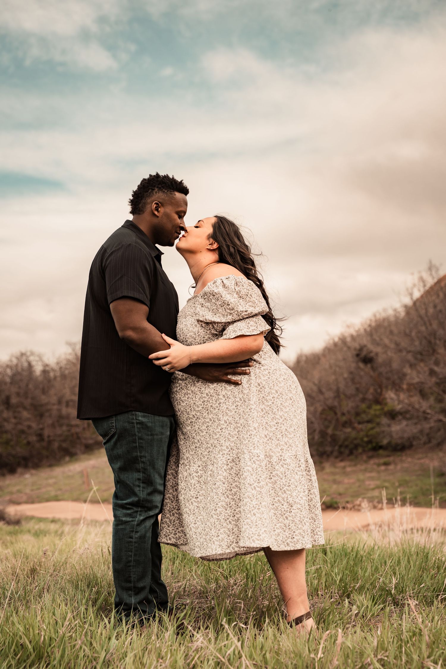 A romantic couple embraces in a grassy field during a cloudy day photo session.