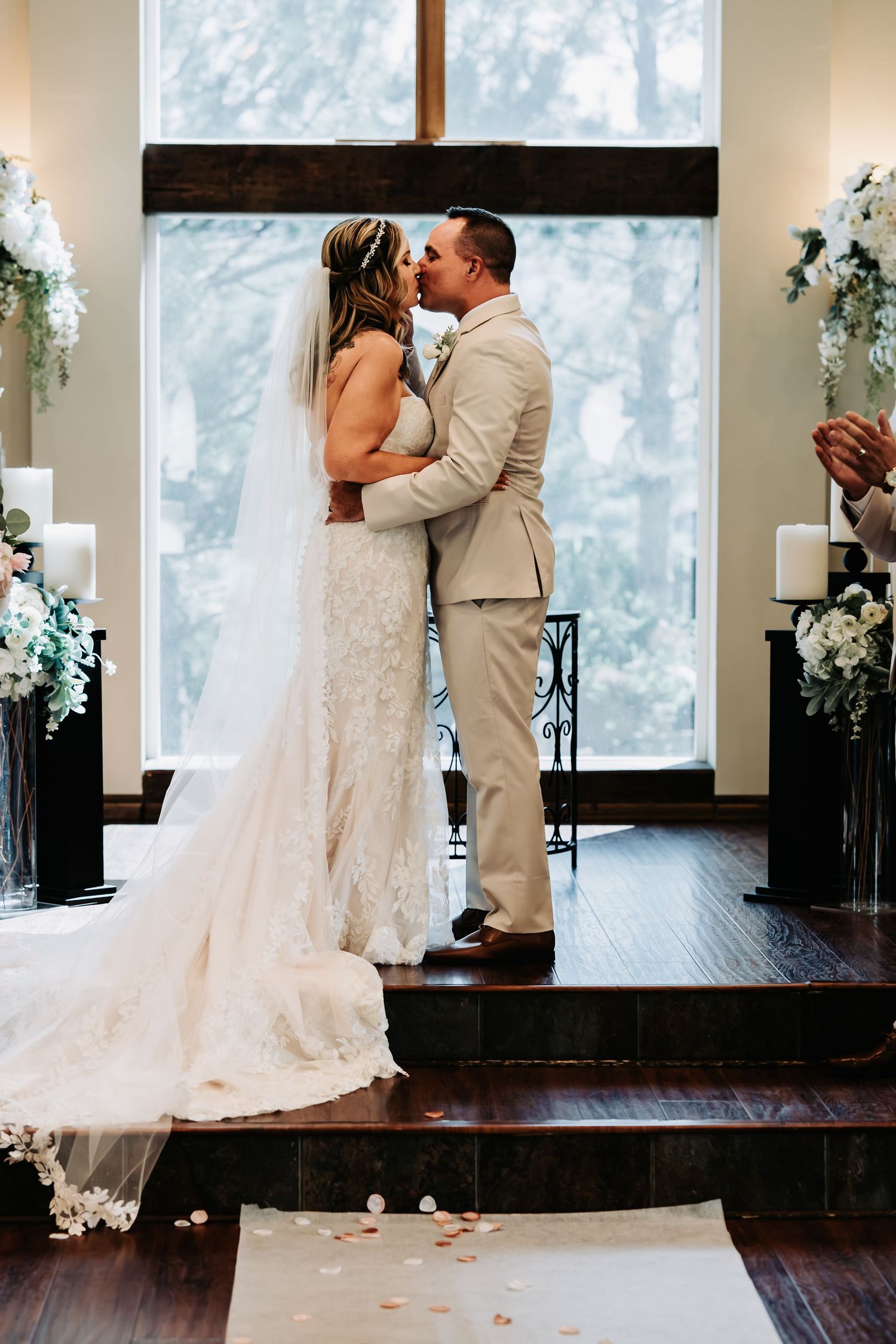 Bride and groom share first kiss at elegant wedding altar adorned with white flowers and candles before a large cross.