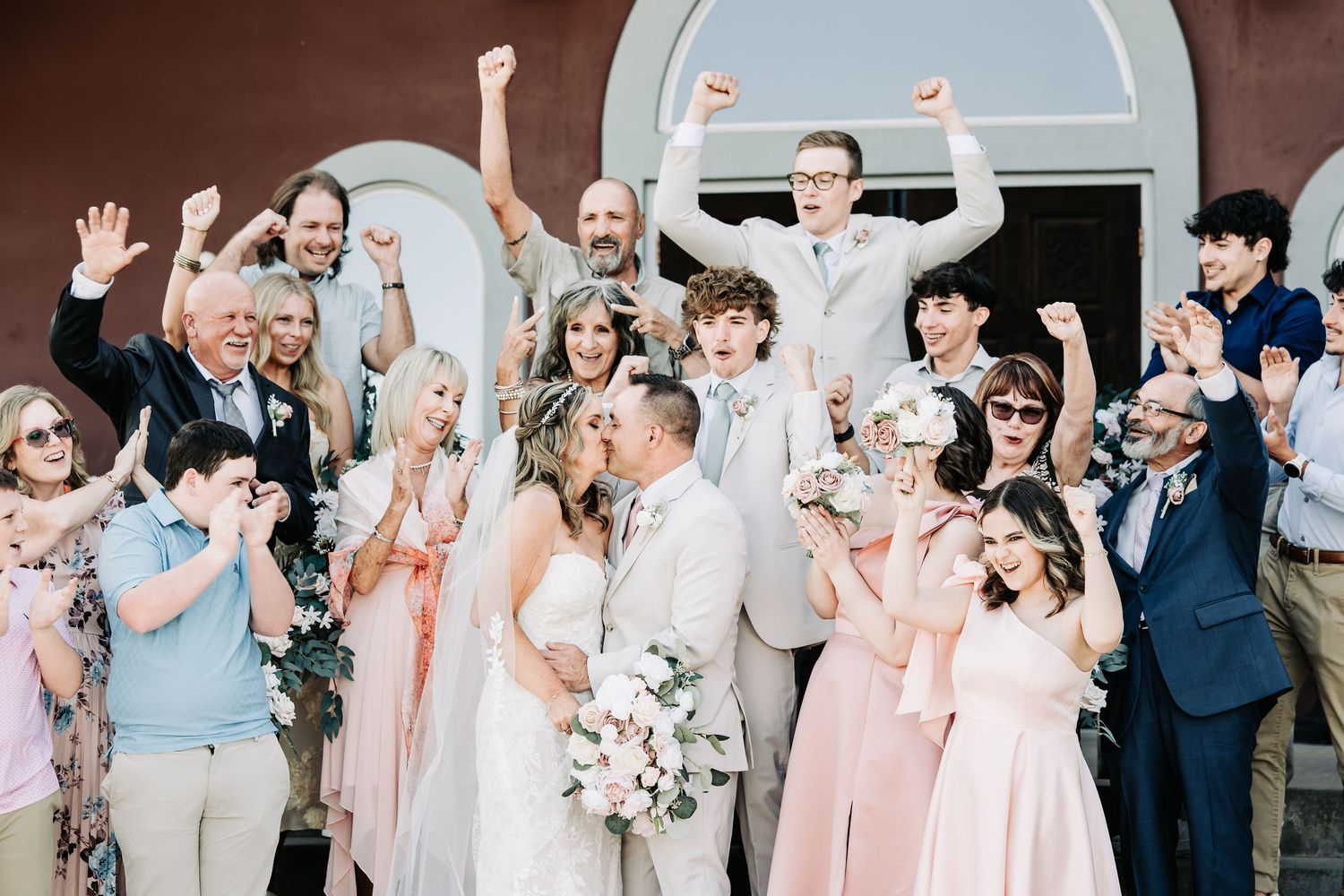 Joyful wedding party celebrating outside a church, bride and groom kissing while guests cheer and throw confetti.