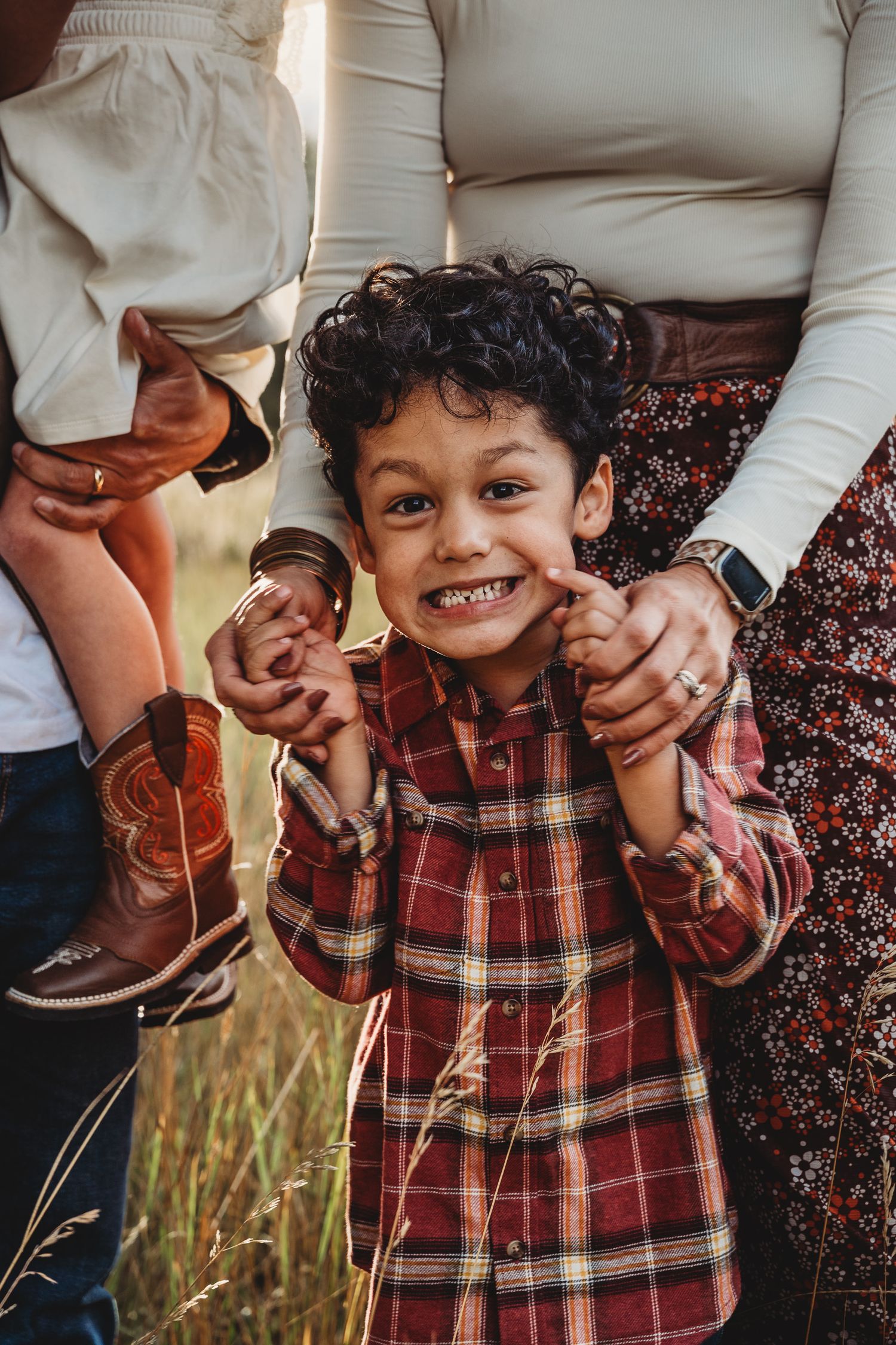 A child wearing a red plaid shirt smiles while holding hands with someone in a floral dress and white top.