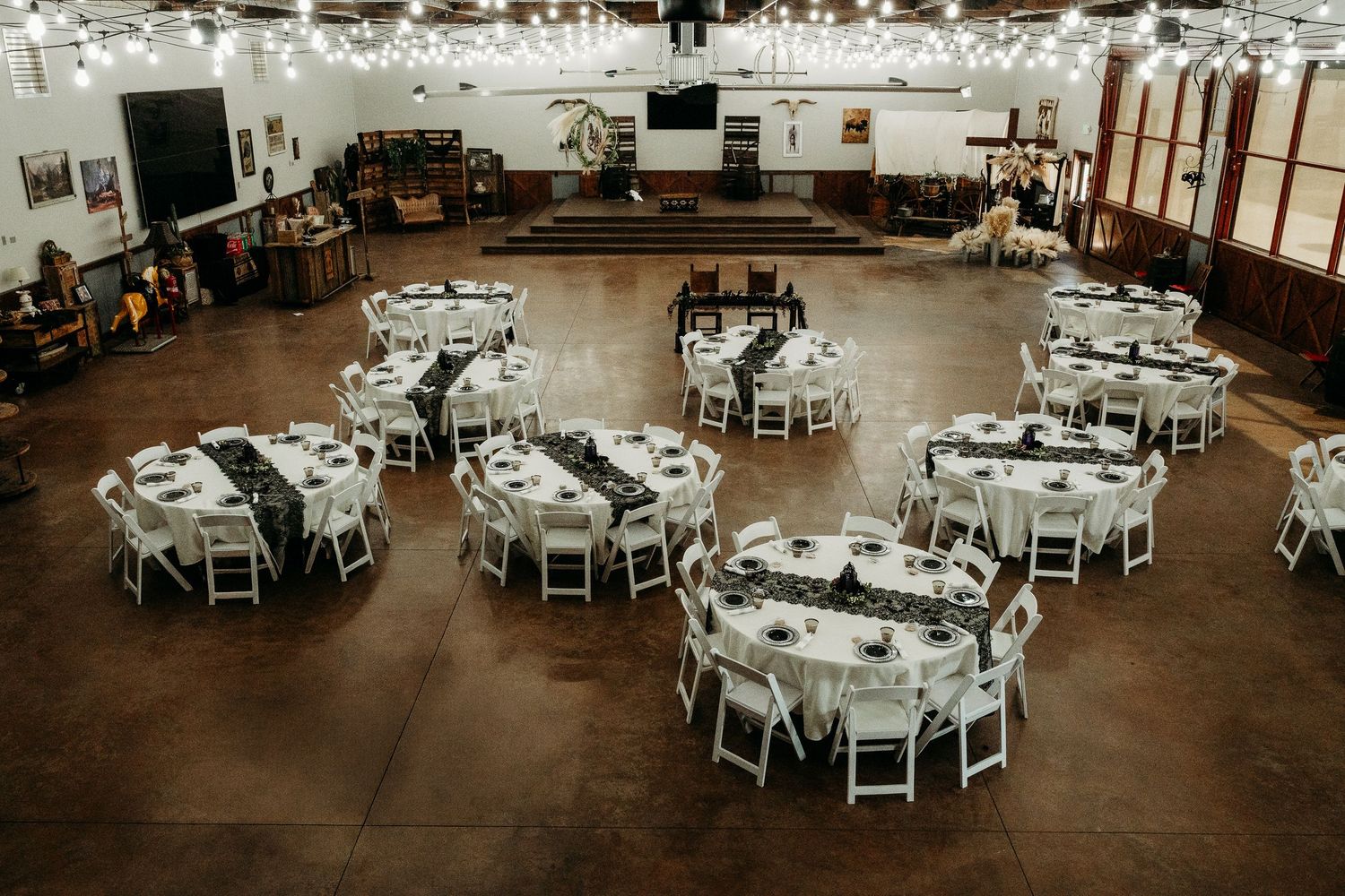 Elegant wedding reception hall with round tables, white chairs, string lights, and dark floral centerpieces on a polished concrete floor.