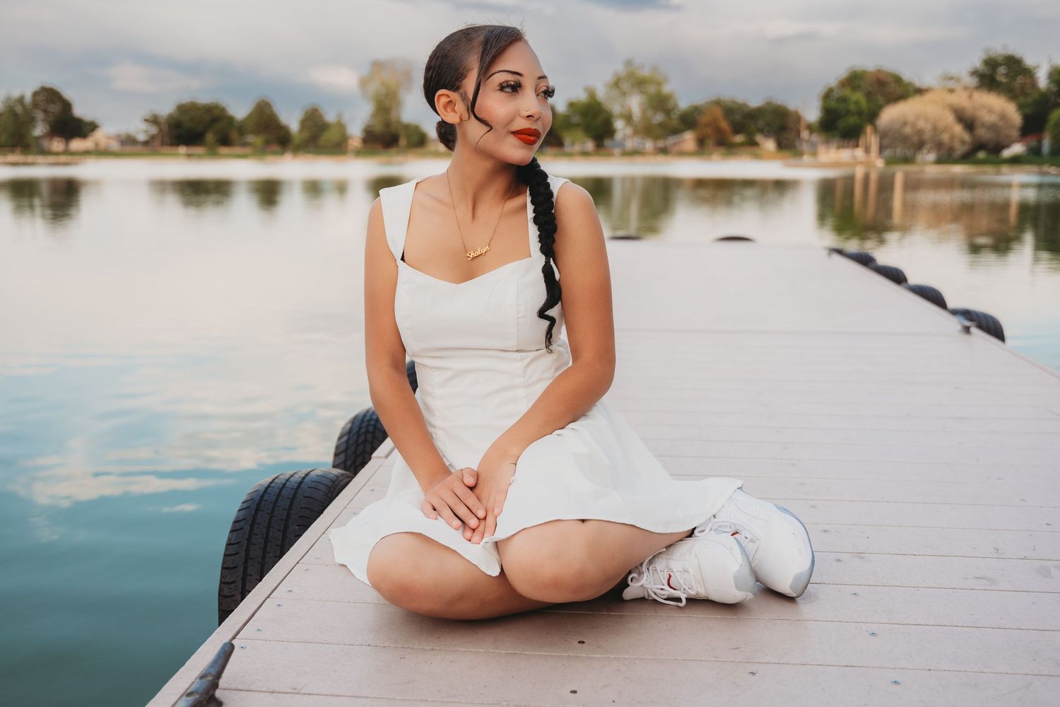 A person in a white dress sits on a dock by a tranquil lake while birds gather nearby.