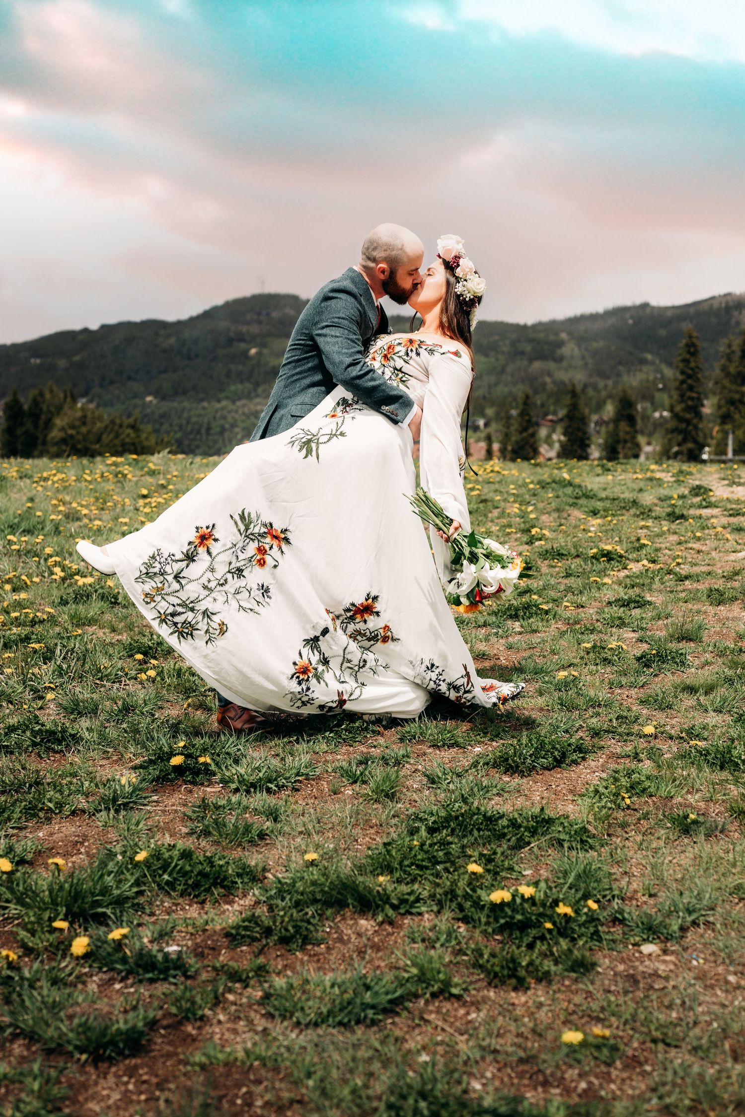 Bride and groom sharing a romantic kiss in a mountain meadow, bride wearing a floral embroidered white wedding dress.