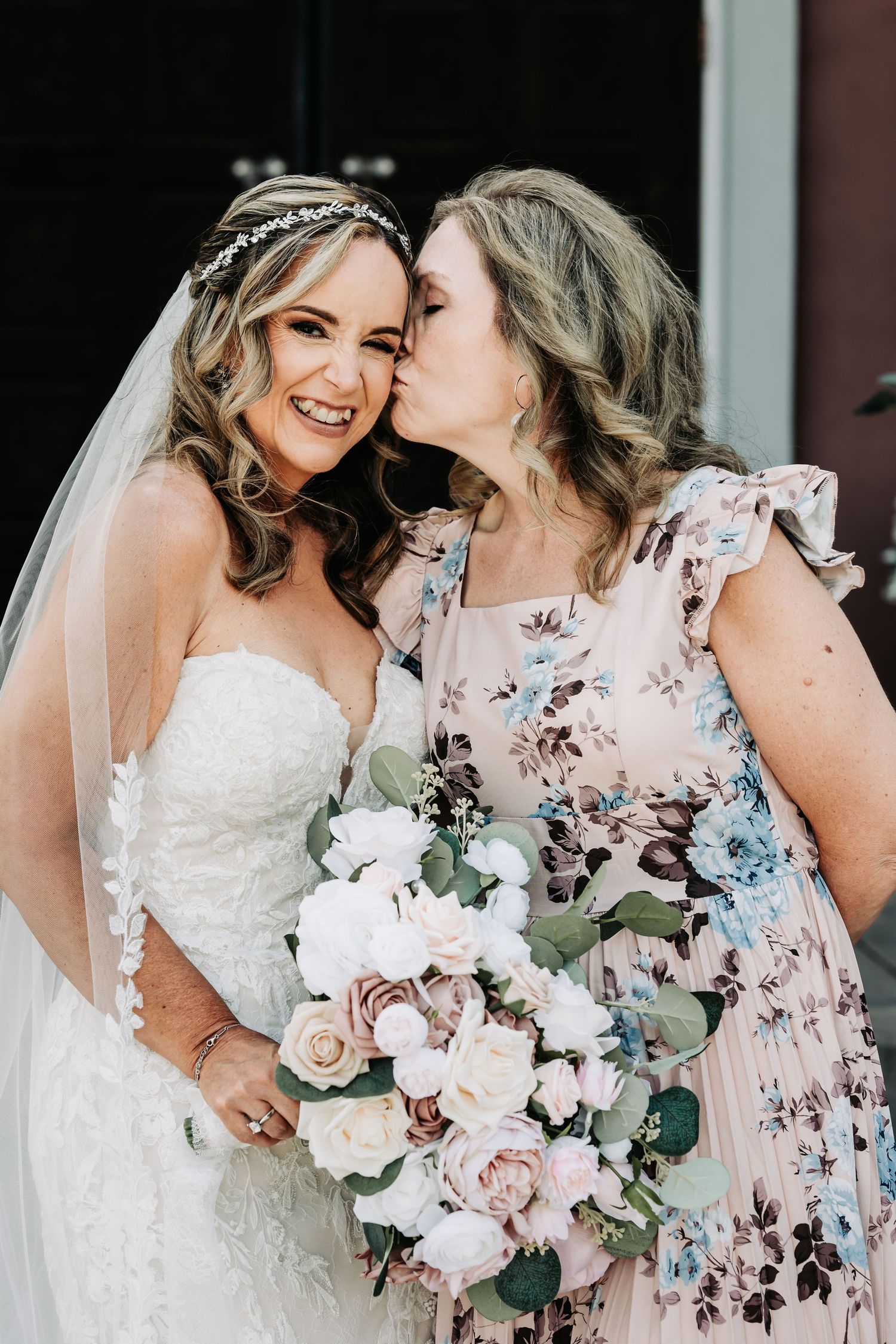 Bride in white lace gown and veil holds bouquet as woman in floral dress kisses her cheek.