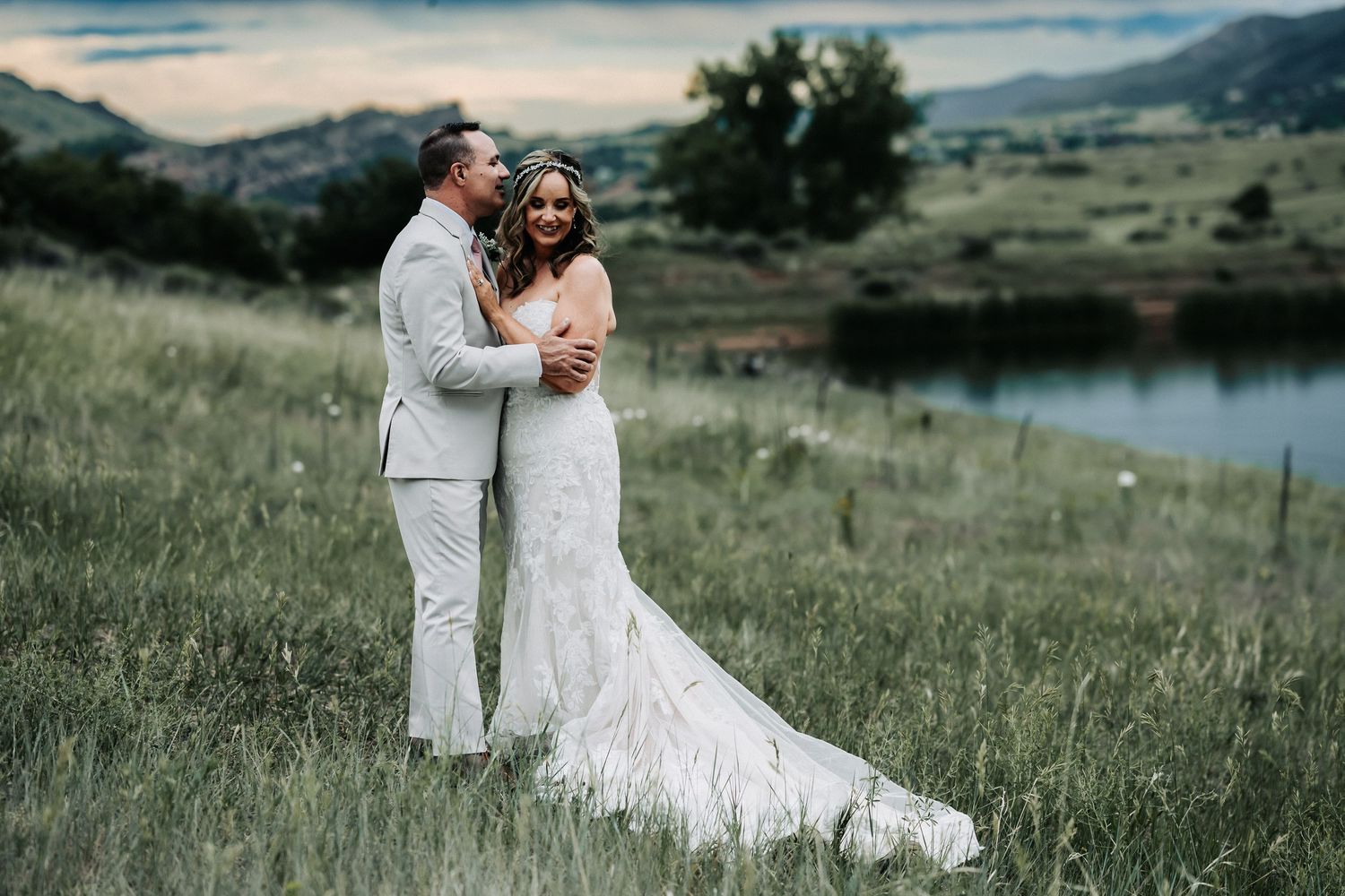 Bride and groom embracing in a lush green meadow with a scenic mountain lake backdrop at golden hour.
