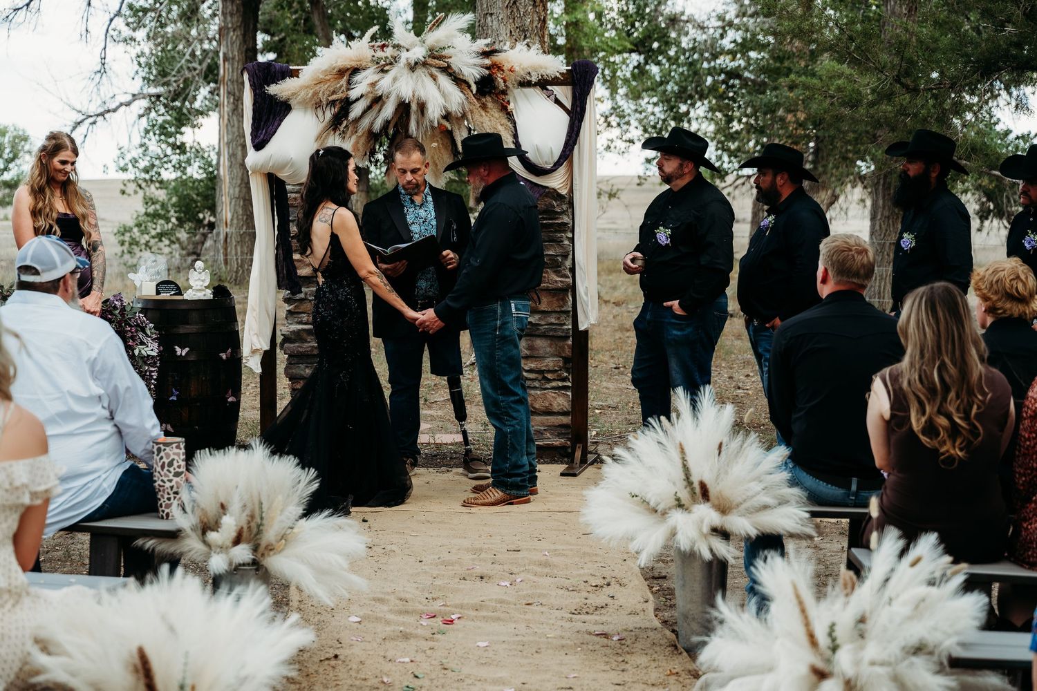 Outdoor wedding ceremony with couple exchanging vows under pampas grass arch, guests in black attire, rustic desert setting.