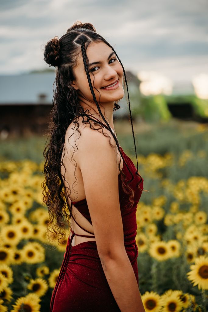 Young woman in red dress smiling in sunflower field with braided curly hair at dusk.