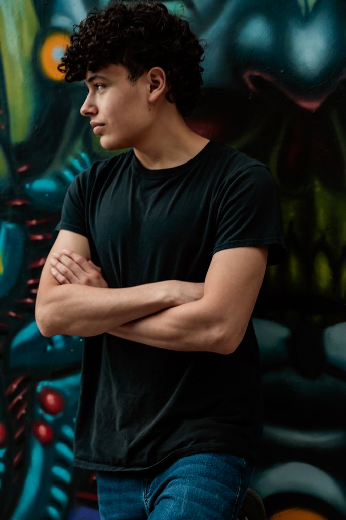 Person wearing black t-shirt poses with crossed arms in moody urban lighting.