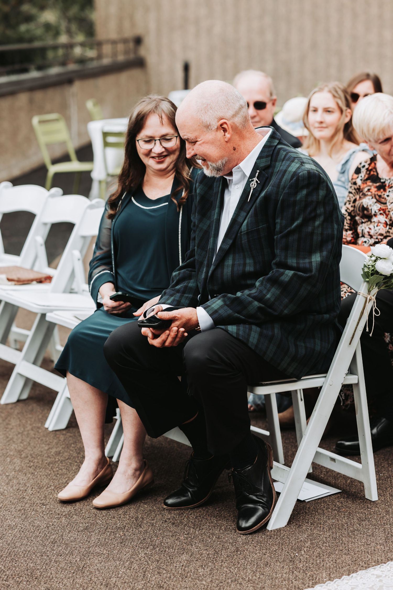 A bald man in plaid jacket and woman in green dress share a warm moment seated at an outdoor wedding ceremony.