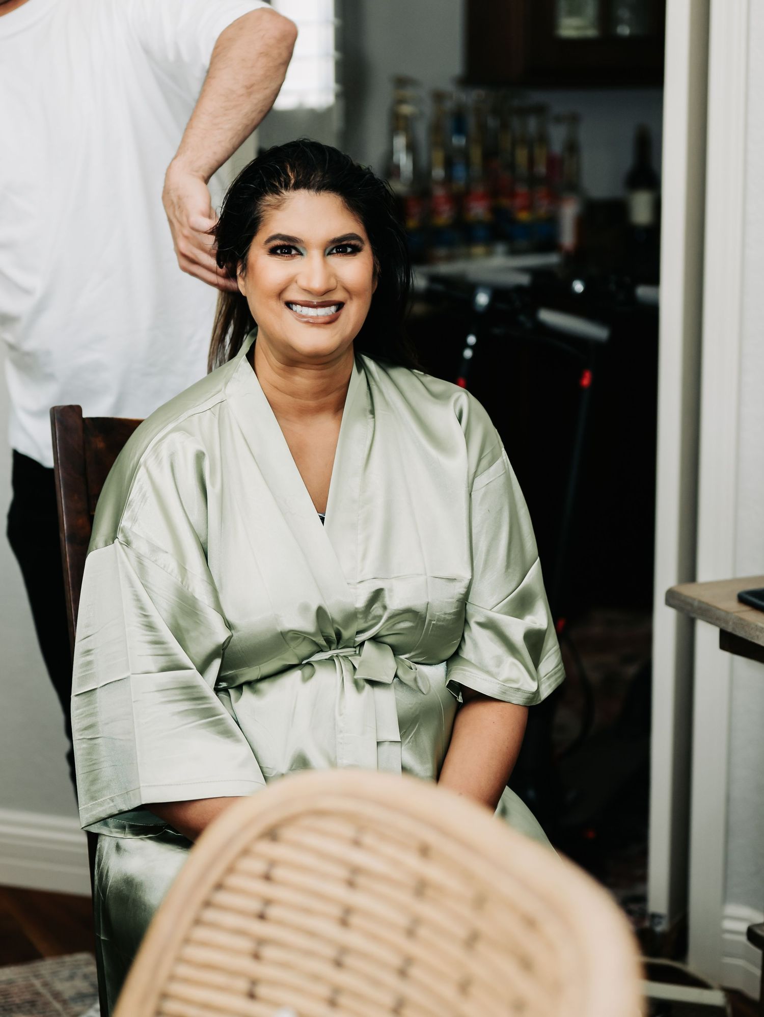 Woman in sage green satin robe smiling while getting hair done during bridal preparation.