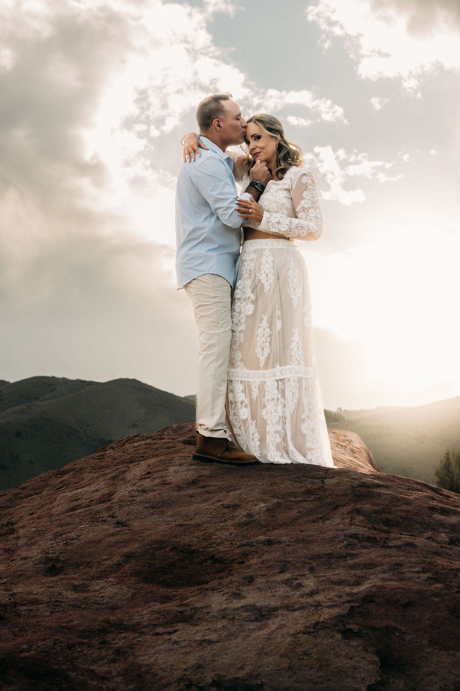 Couple in wedding attire embracing on a rocky mountain summit at golden hour with dramatic cloudy sky backdrop.