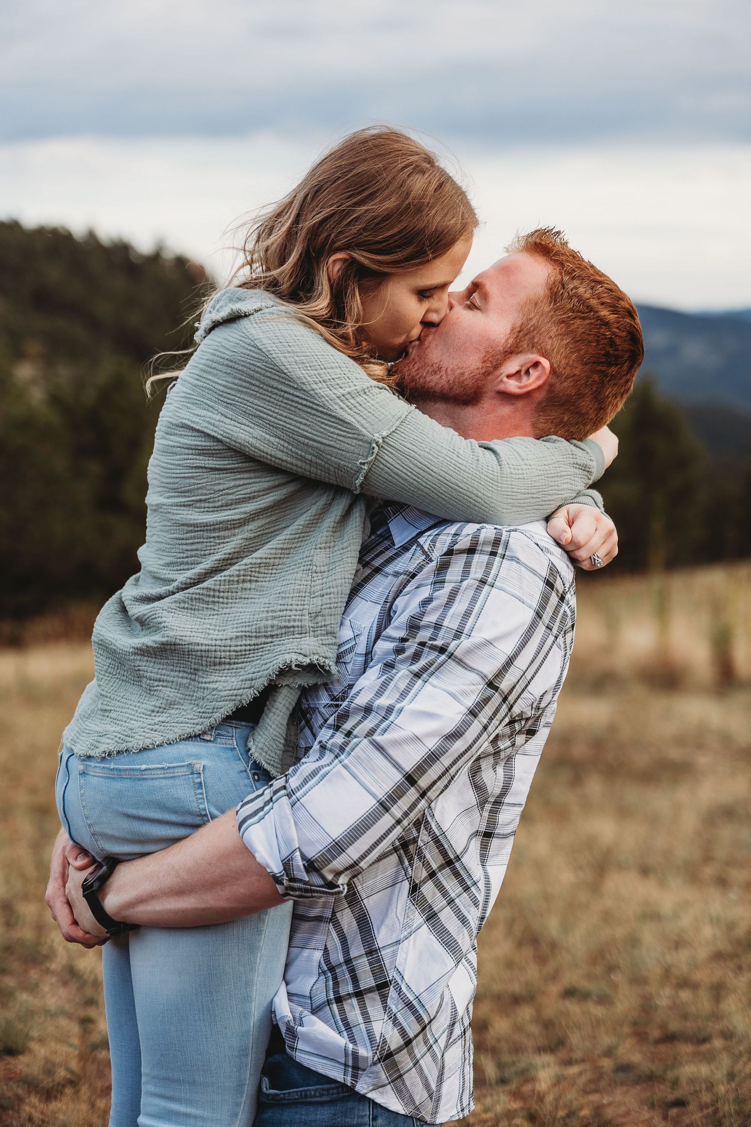 A couple embraces in a tender moment against a mountainous backdrop with golden grass and cloudy skies.