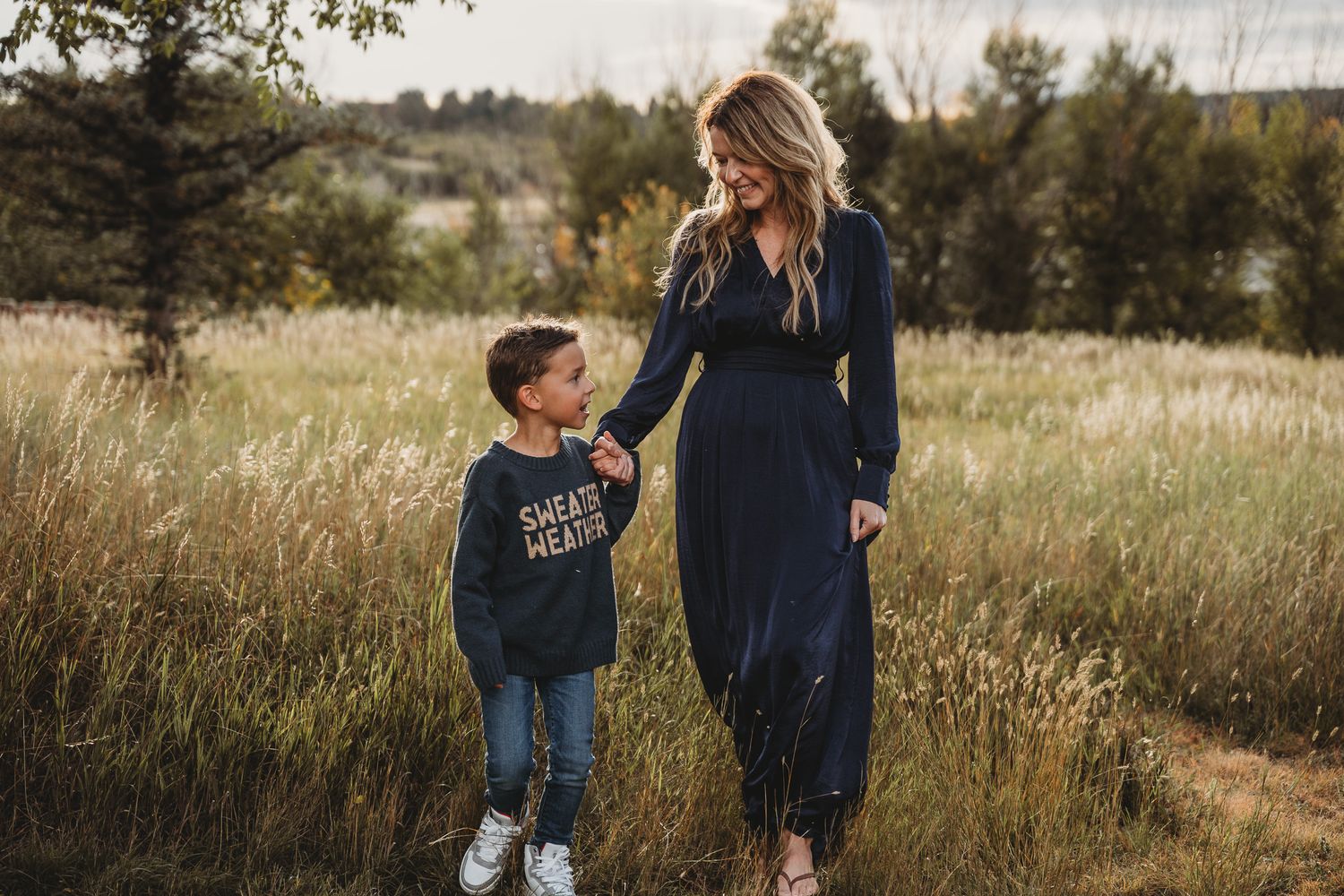 Mother and child walk together on a winding dirt path through a scenic autumn meadow at sunset.