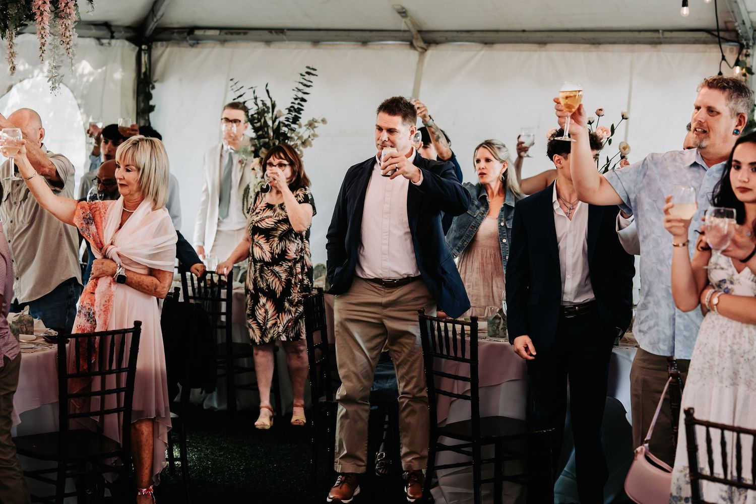 Guests raising glasses in a toast at an elegant outdoor wedding reception under a white tent.