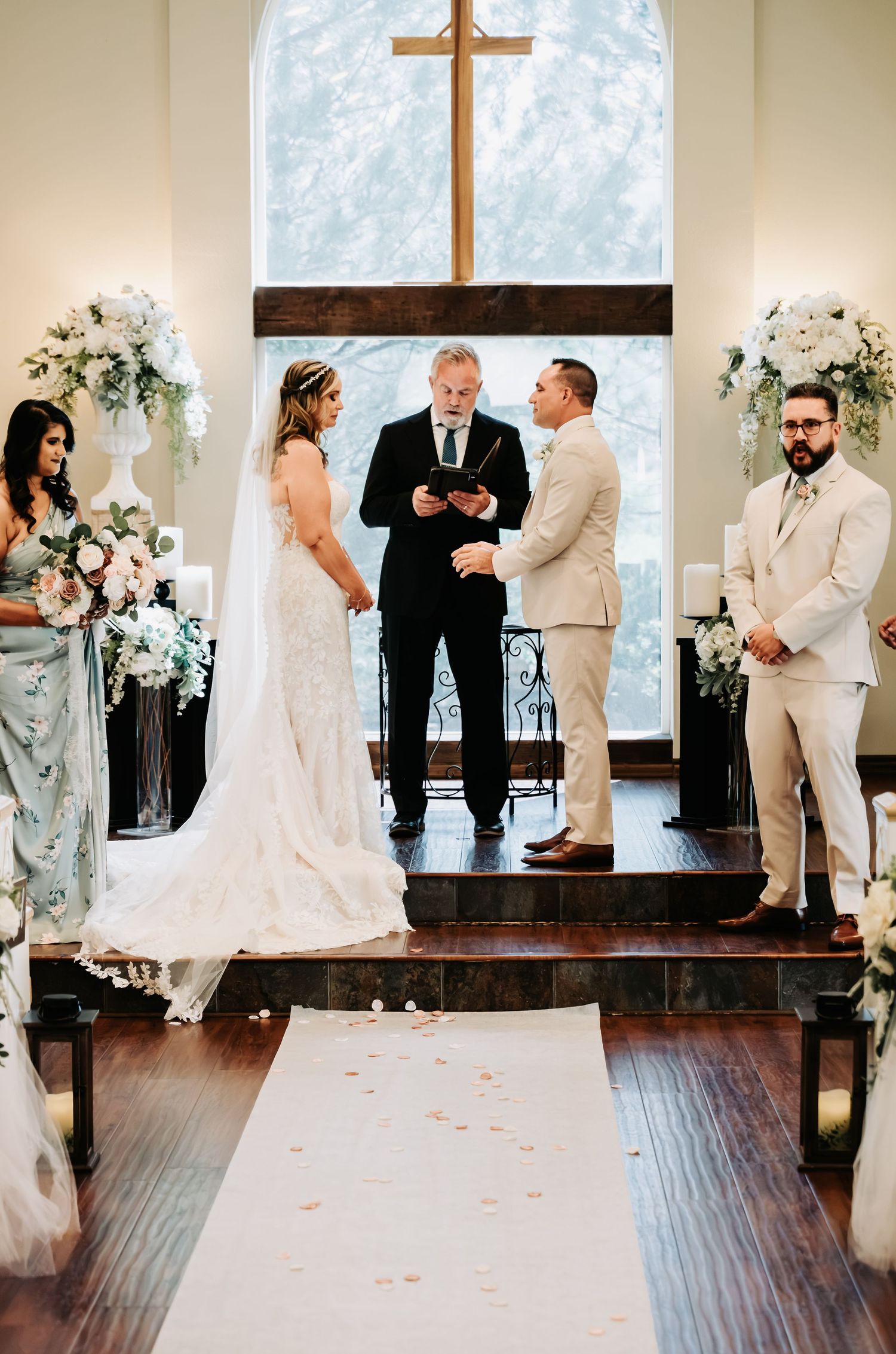 Bride and groom exchange vows at elegant chapel altar with white floral arrangements and wooden cross backdrop.