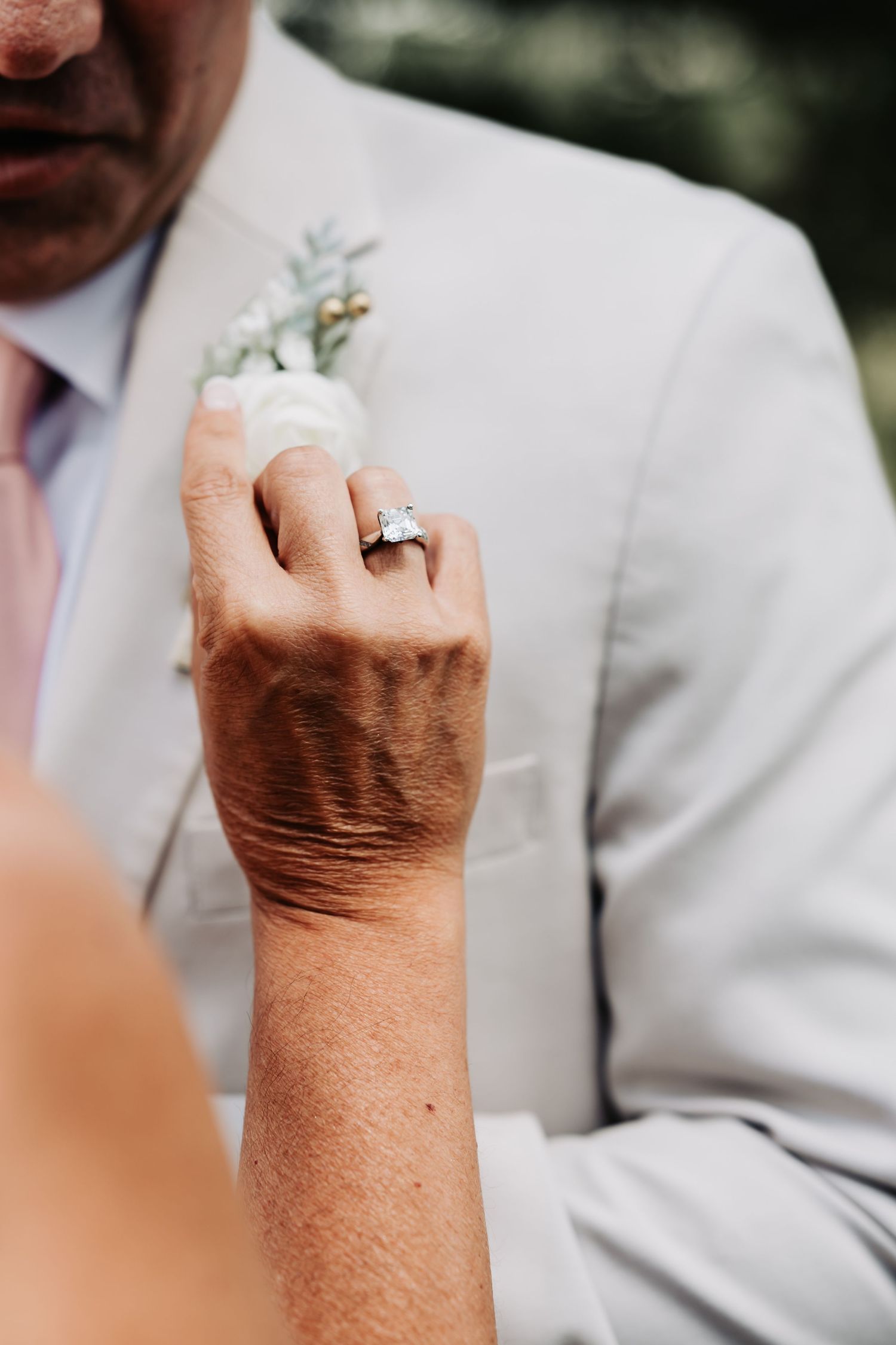 Close-up of a bride's hand with a diamond engagement ring adjusting a boutonniere on a groom's white suit lapel.