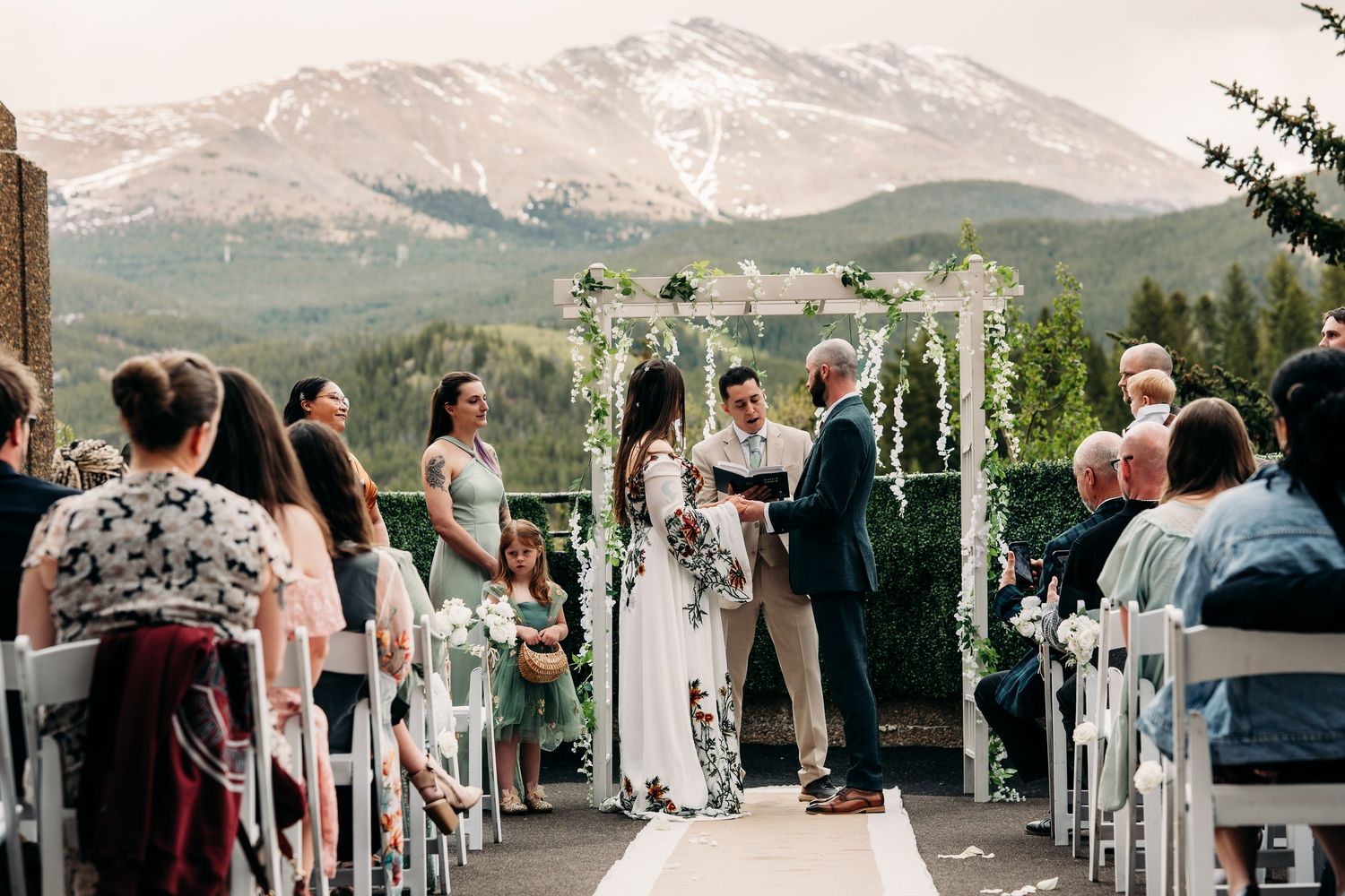 Outdoor mountain wedding ceremony with floral arch, guests seated in white chairs, and snow-capped peaks in the background.