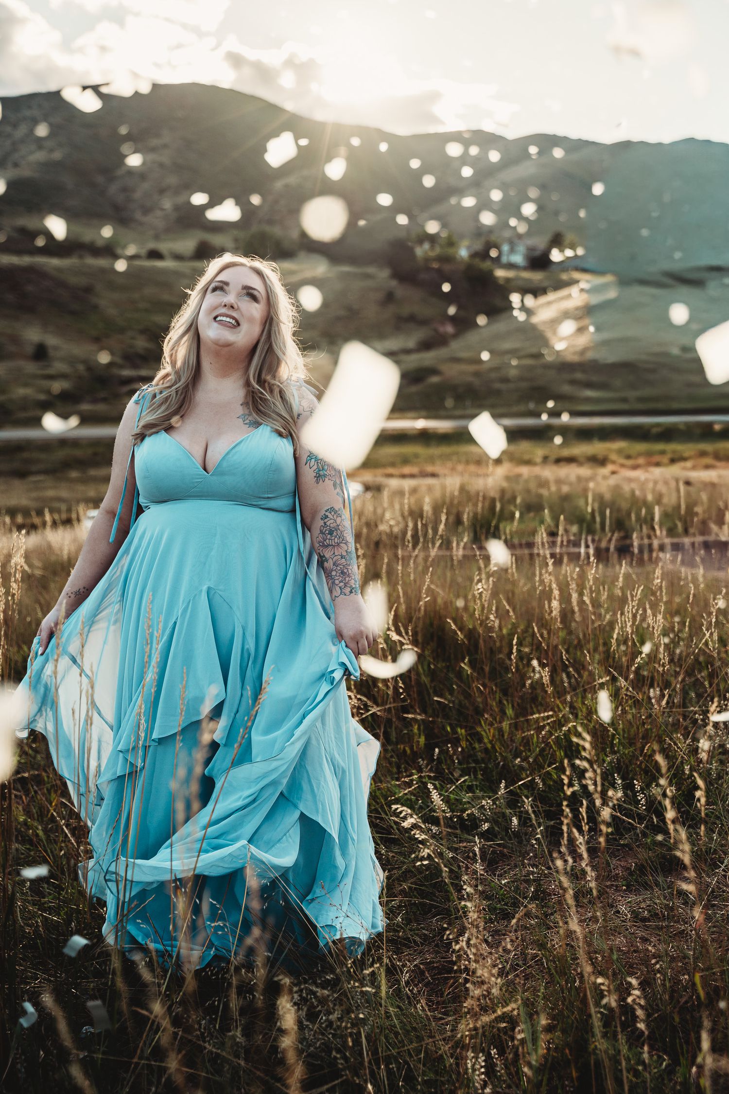A person in a flowing turquoise dress stands in a sunlit meadow with floating white bokeh effects in the air.
