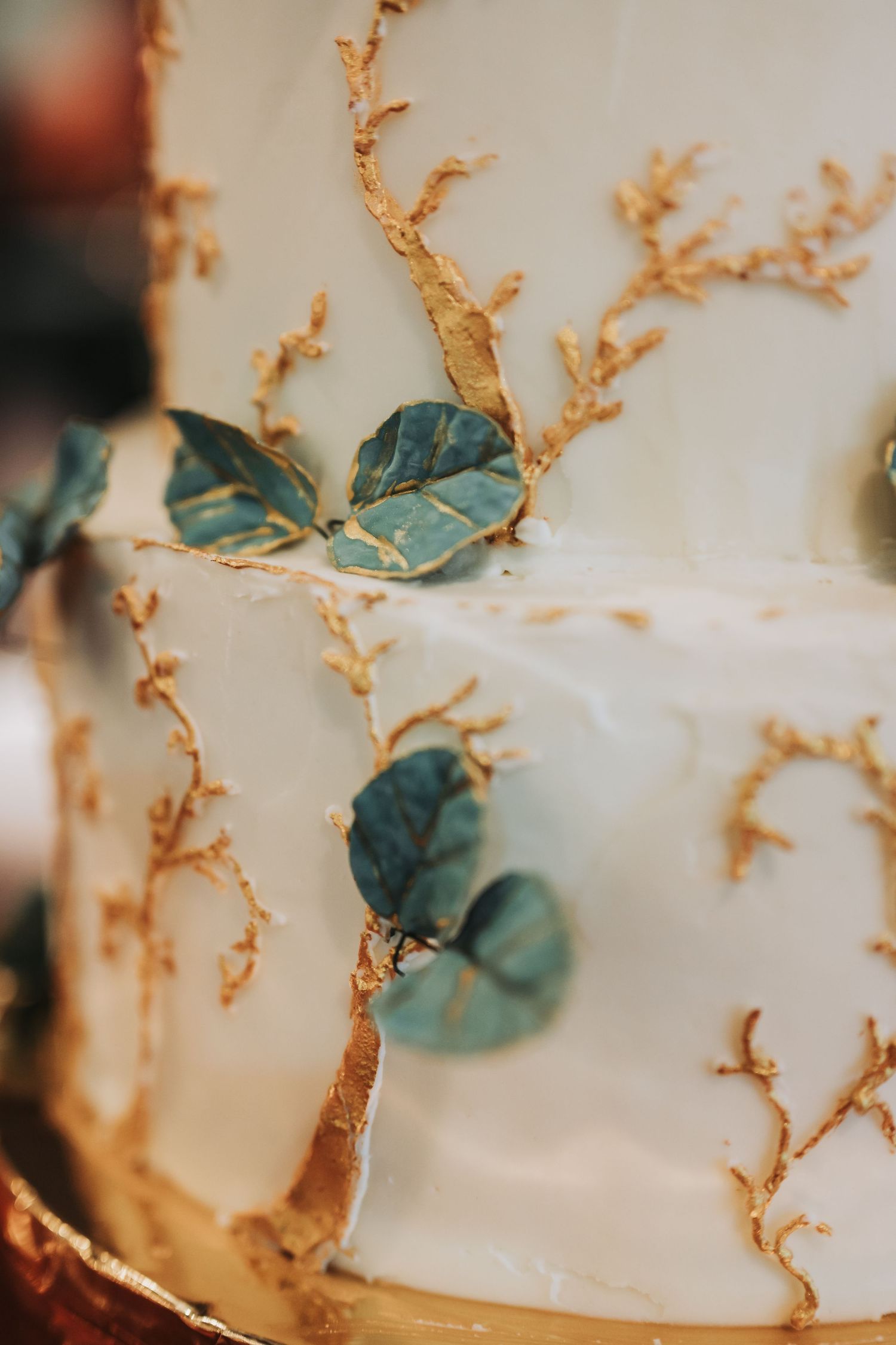 Elegant white wedding cake with gold branch details and teal eucalyptus leaves decoration.