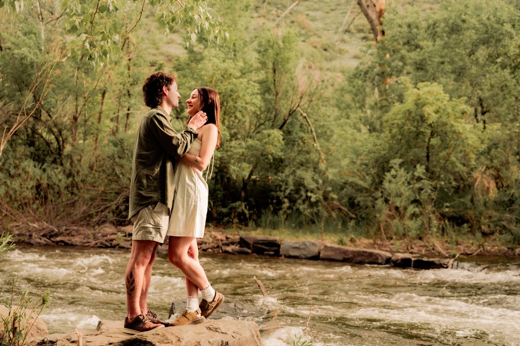 A romantic couple shares a tender moment dancing together on a sandy riverbank surrounded by trees.