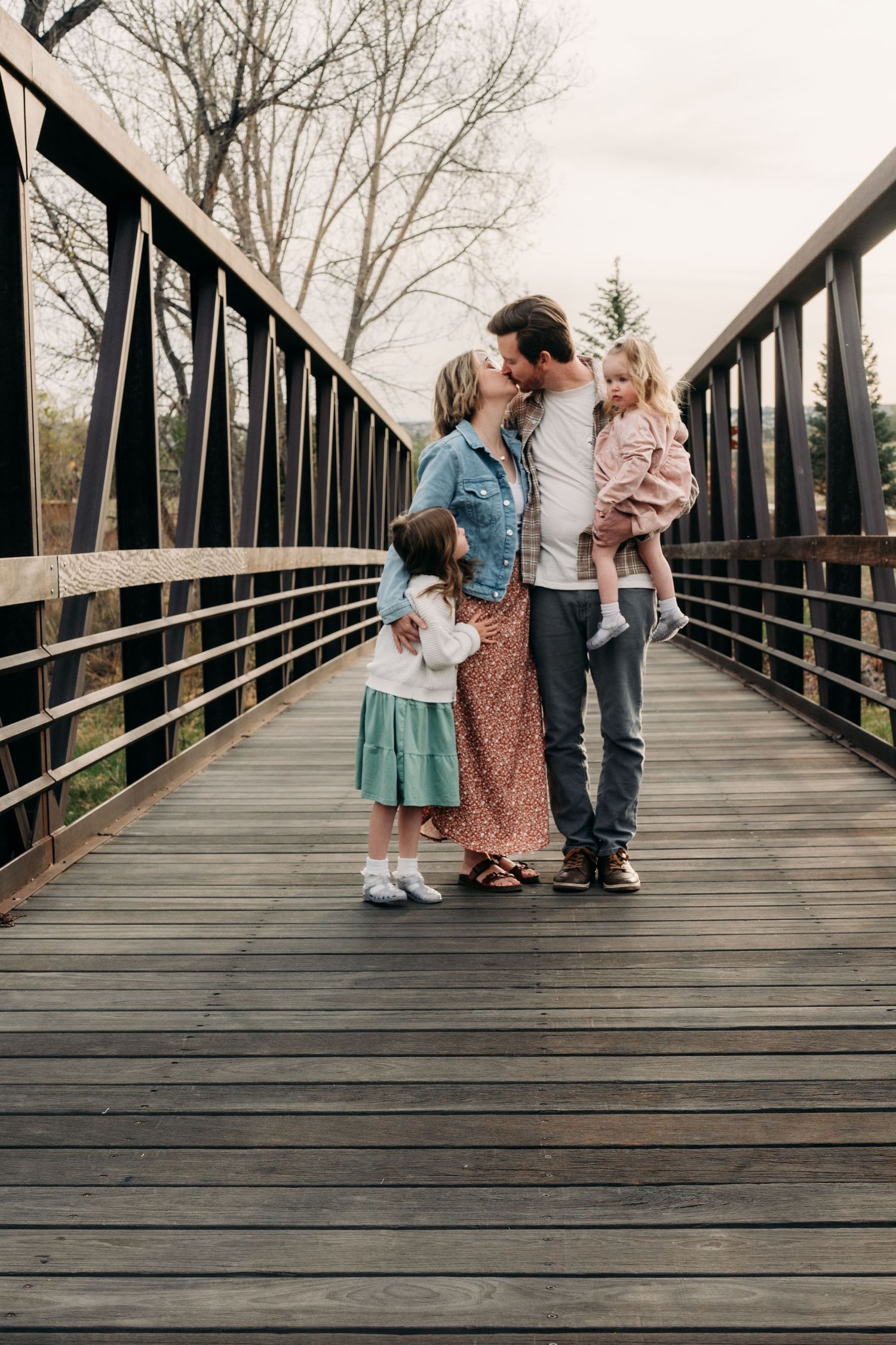 Family of four sharing a tender kiss on a wooden bridge, with two young girls in spring attire.