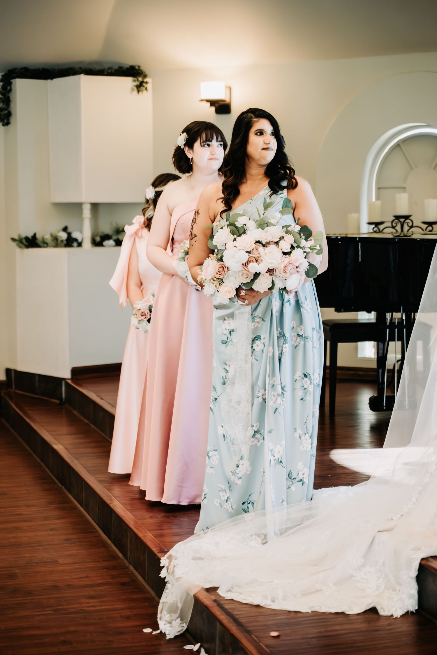 Bridesmaids in pink and floral blue dresses stand at wedding altar holding bouquets of soft pink flowers.