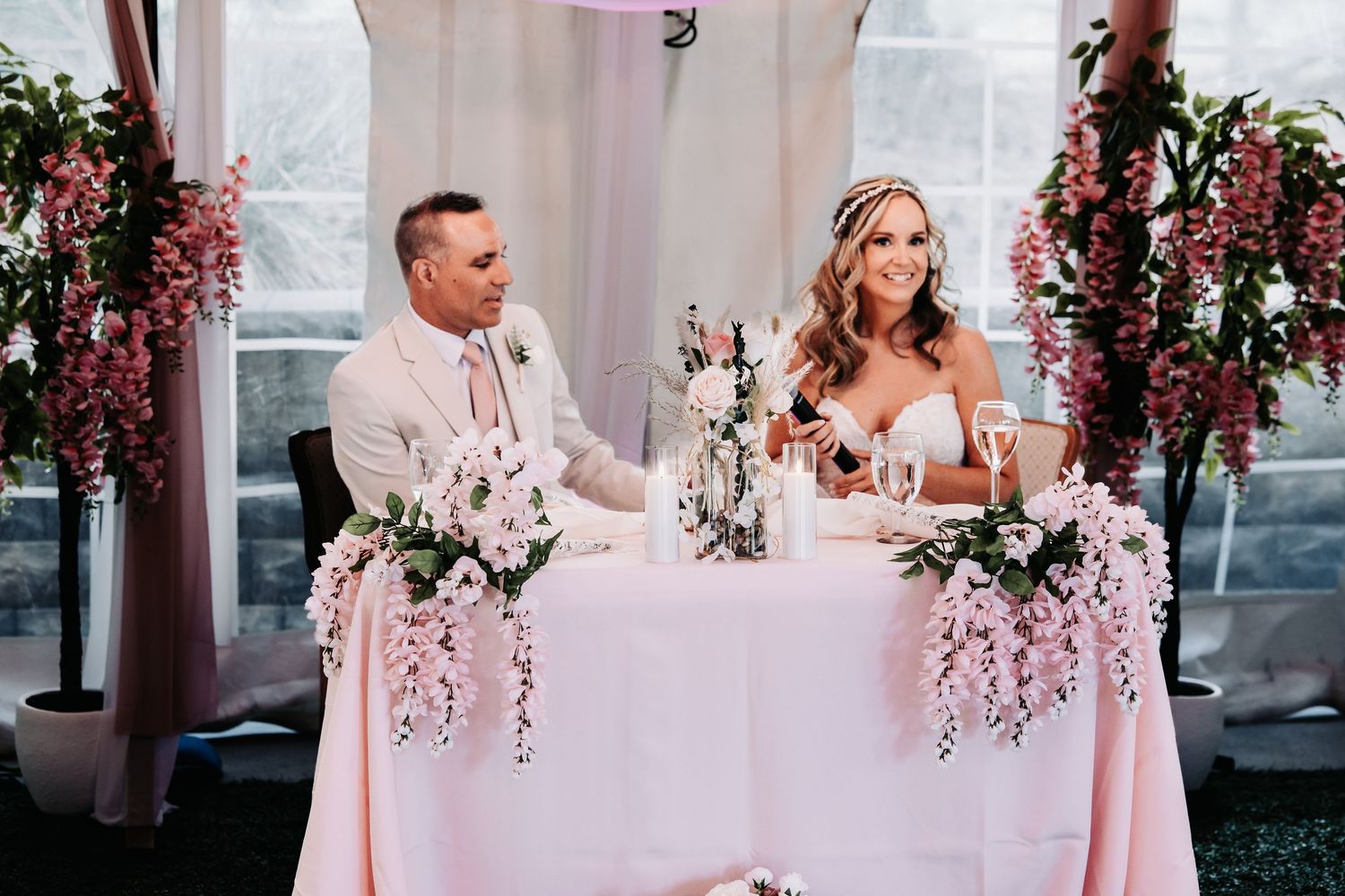 Bride and groom smiling at elegant sweetheart table adorned with cascading pink flowers at wedding reception.