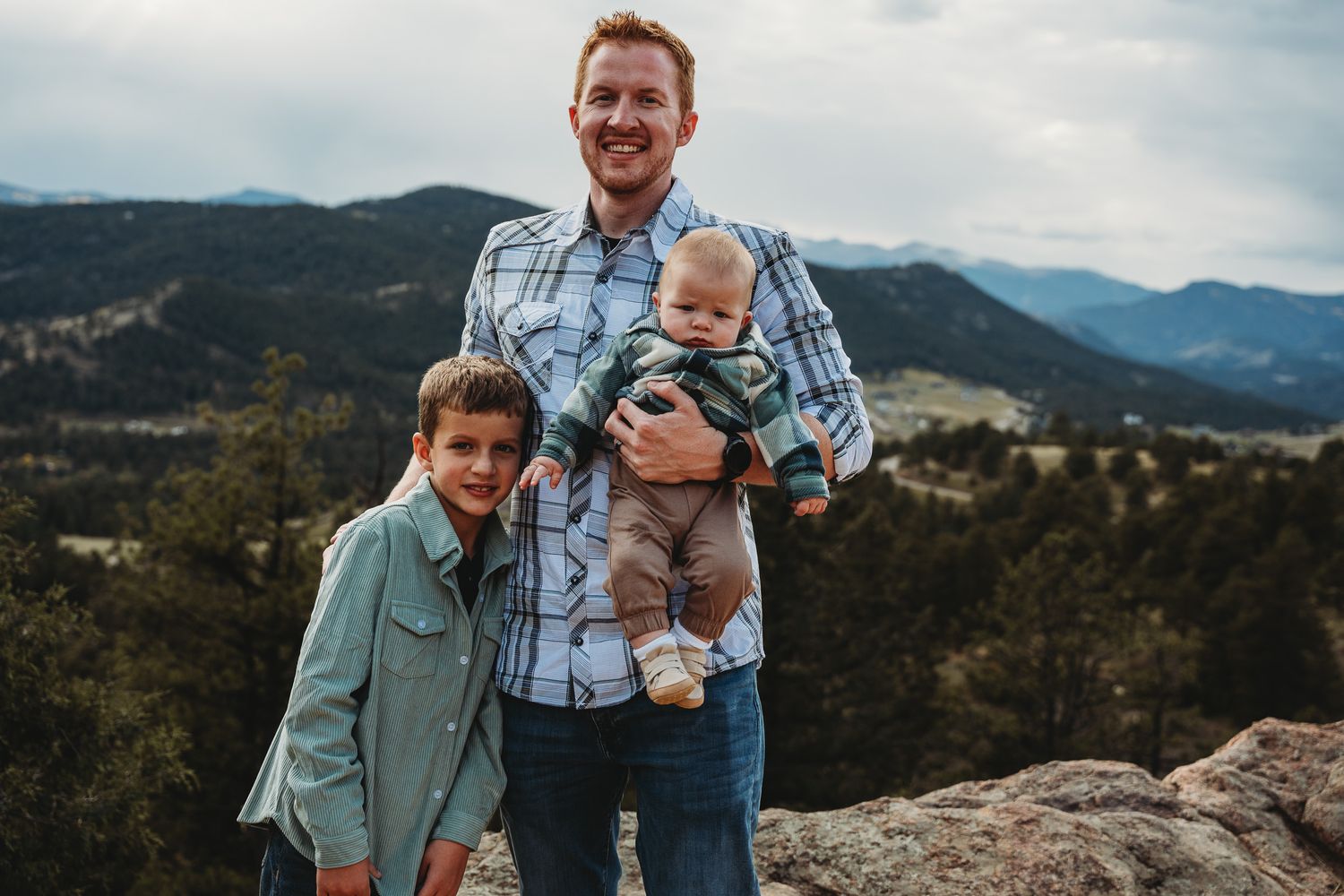 A family stands together on a rocky mountain overlook with scenic mountain ranges in the background.