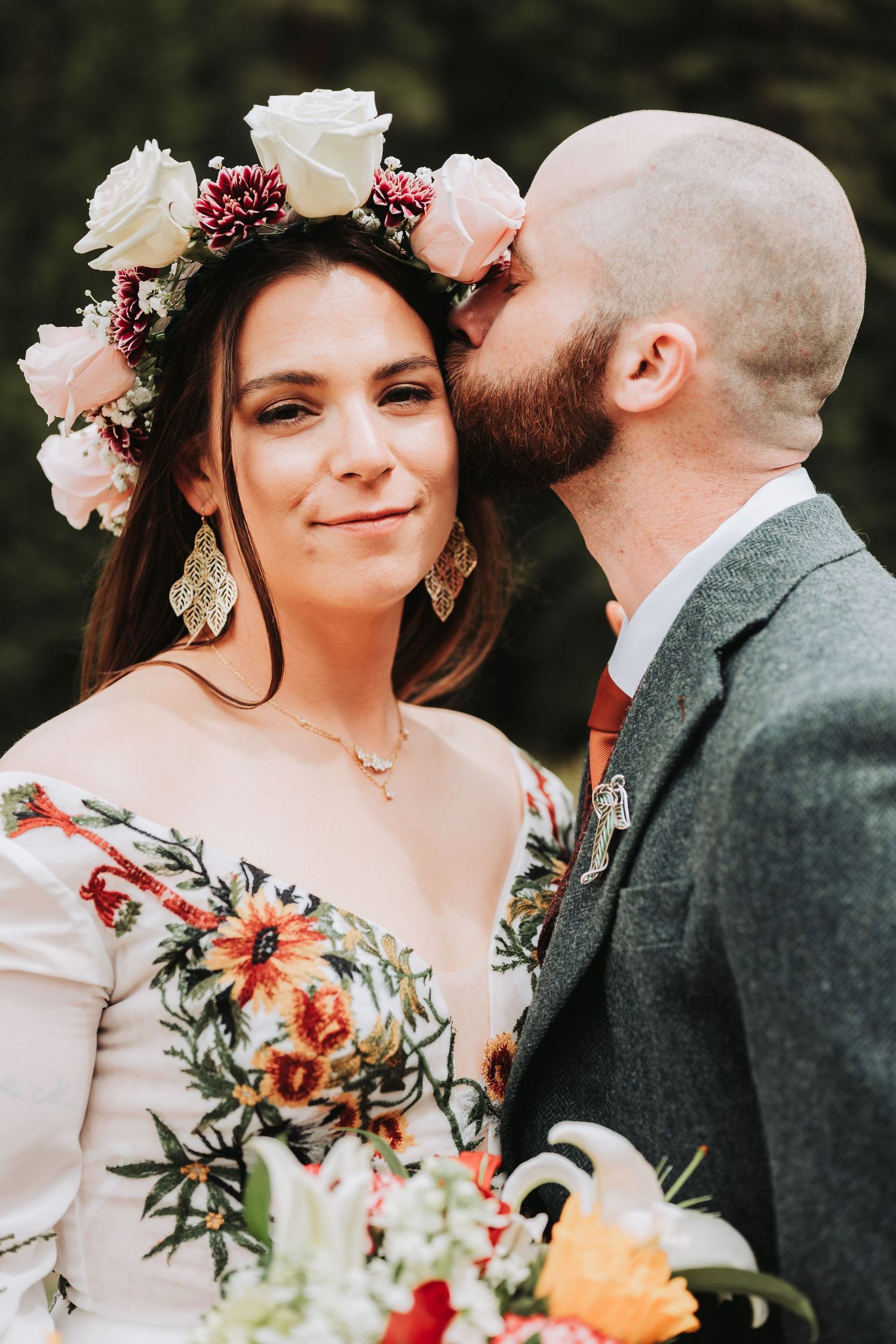 A groom in a gray suit kisses his bride wearing a floral crown and embroidered dress on their wedding day.