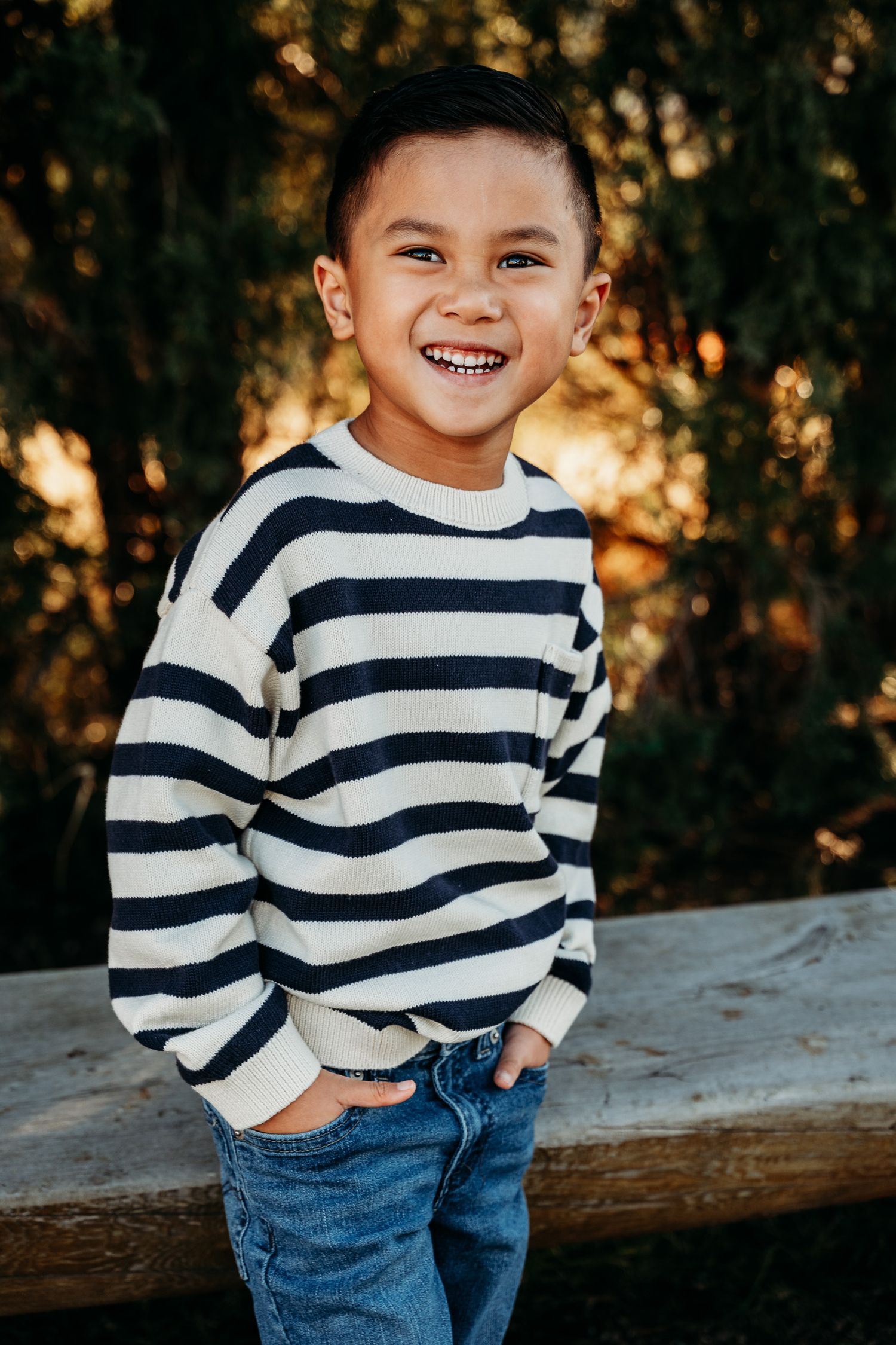 A young child wearing a navy striped sweater and jeans smiles outdoors during golden hour.