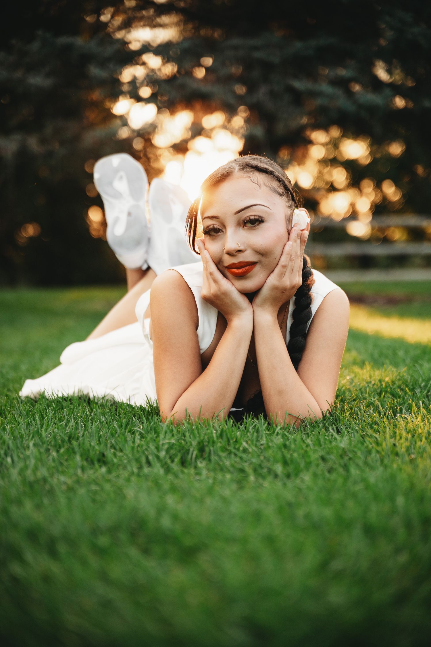Young woman with braids in white dress lying on green grass, smiling at camera during golden hour sunset.