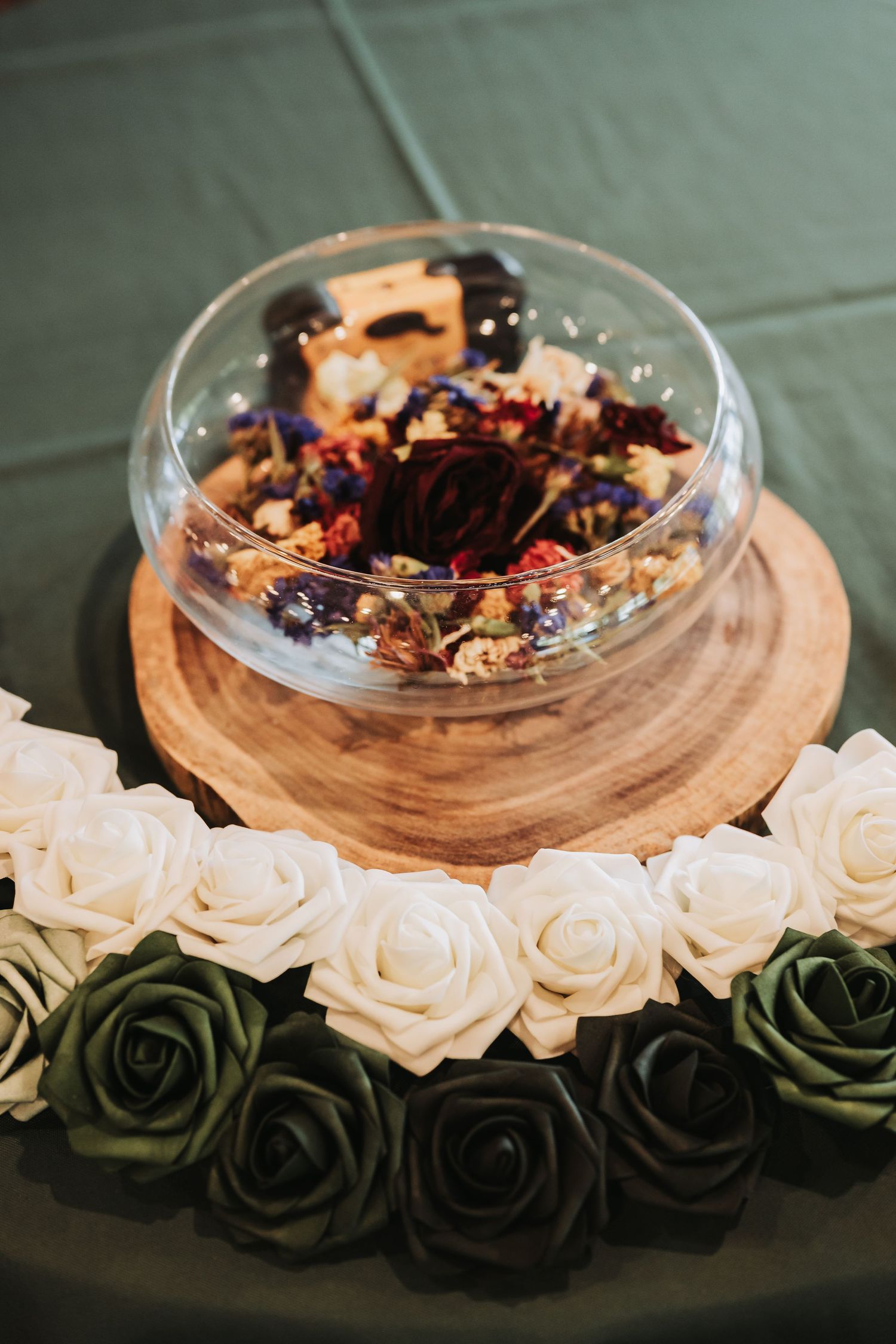 Glass bowl filled with dried flowers and herbs on a wooden board, surrounded by white and green roses on a green tablecloth.
