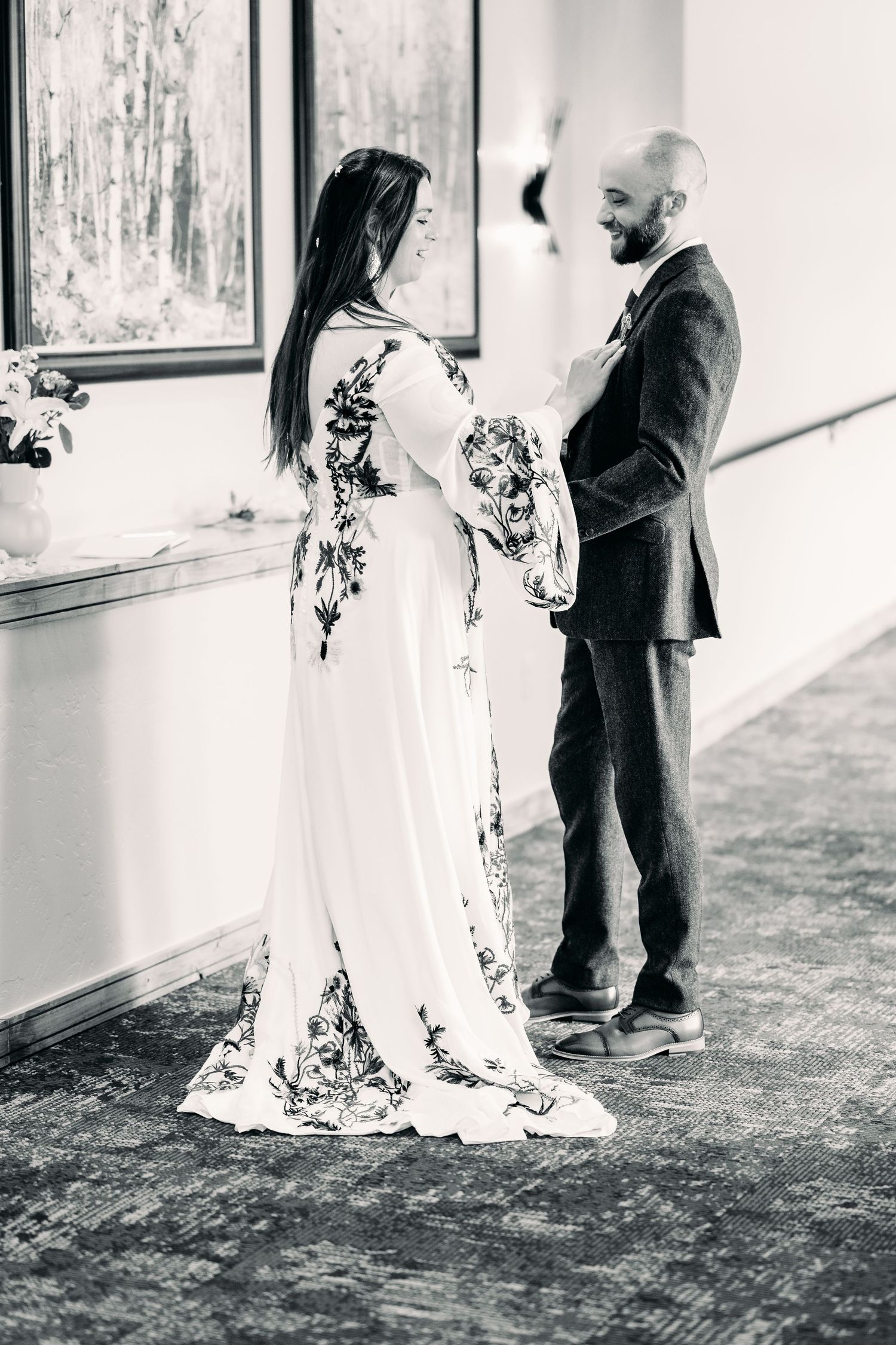 Black and white photo of a bride in floral gown and groom in suit holding hands at an indoor wedding ceremony.