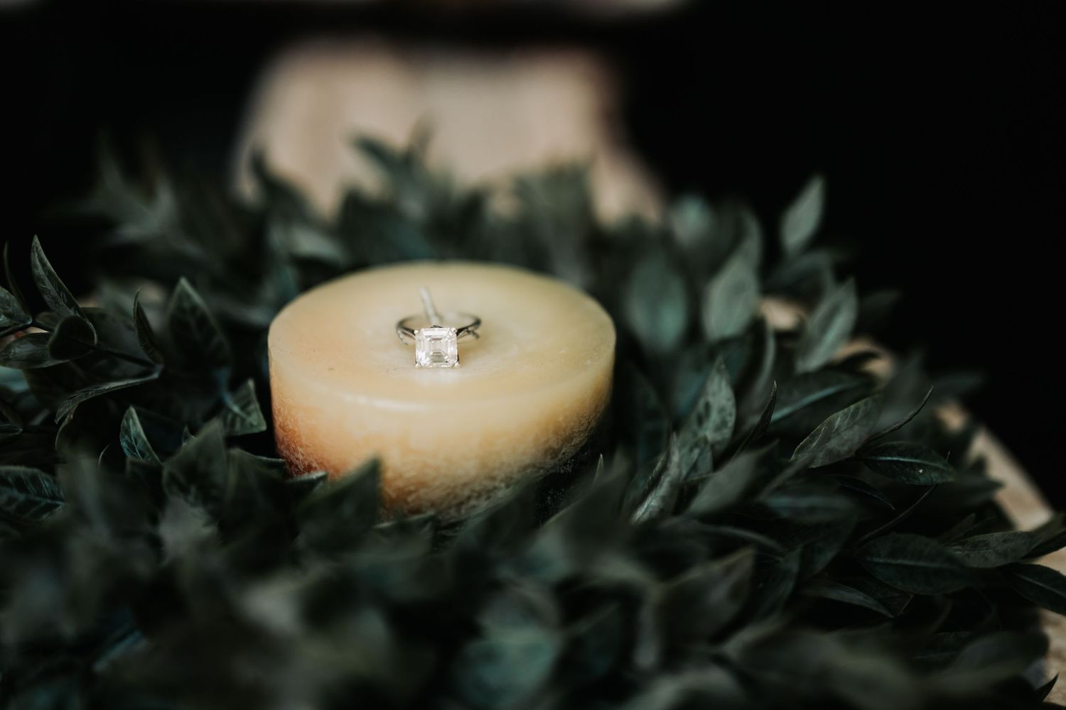 A diamond engagement ring resting on a small amber candle surrounded by lush green leaves.