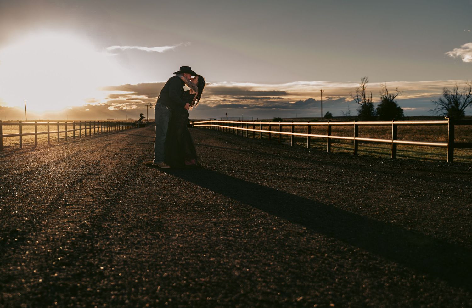 Silhouette of a couple kissing on a gravel road at sunset with wooden fences and golden sky.