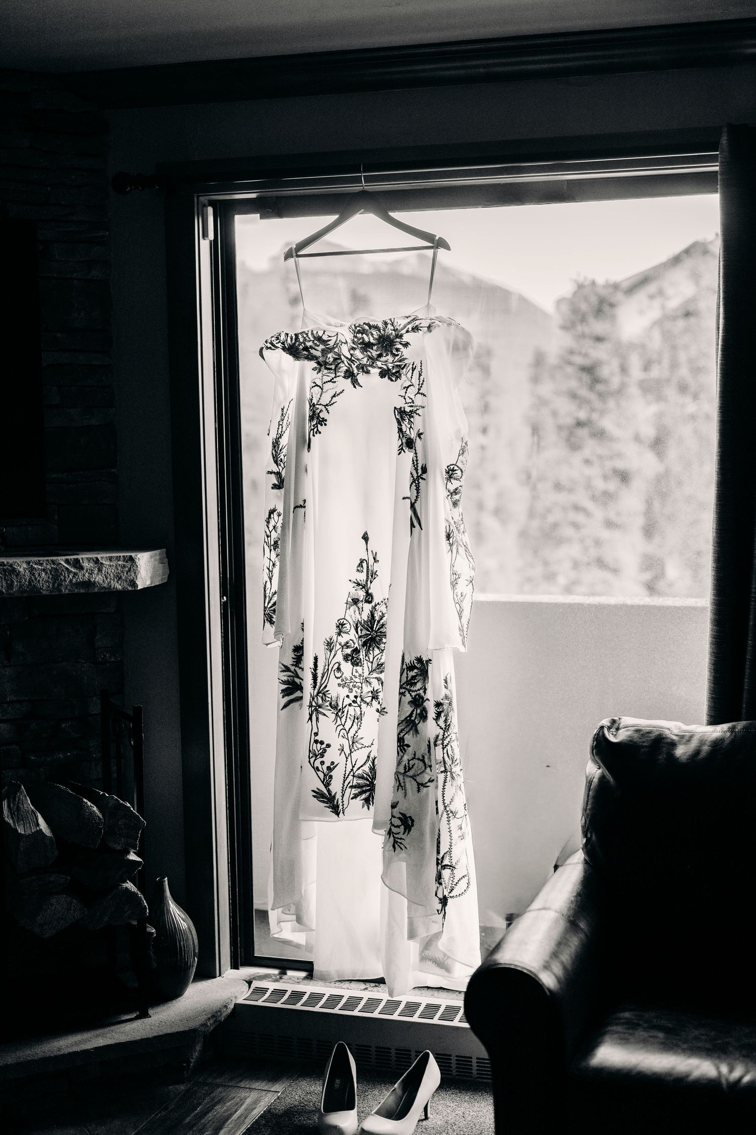 Black and white photo of a floral wedding dress hanging in a window with mountain views and heels below.