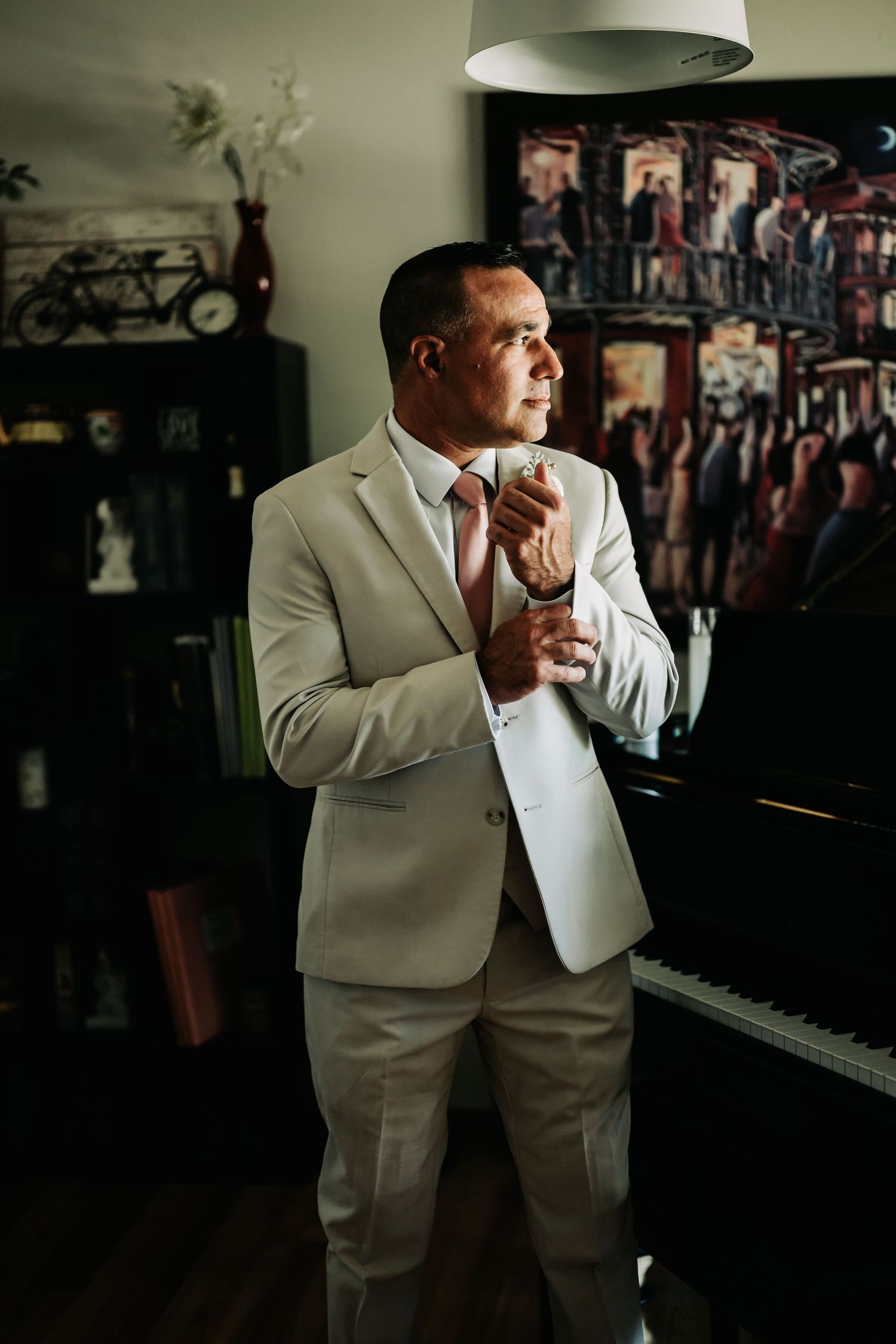 A well-dressed man in a beige suit adjusts his tie while holding a drink near a grand piano indoors.