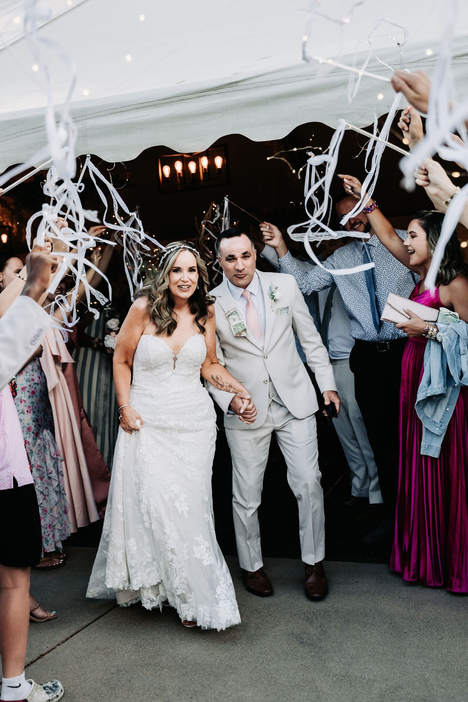 Bride and groom exit wedding reception through ribbon wand tunnel surrounded by cheering guests under a white tent.