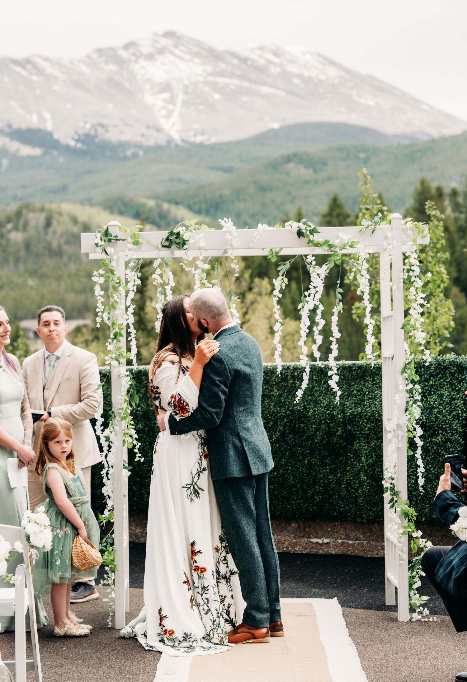Bride and groom share first kiss at outdoor mountain wedding ceremony beneath white floral arch in Colorado.