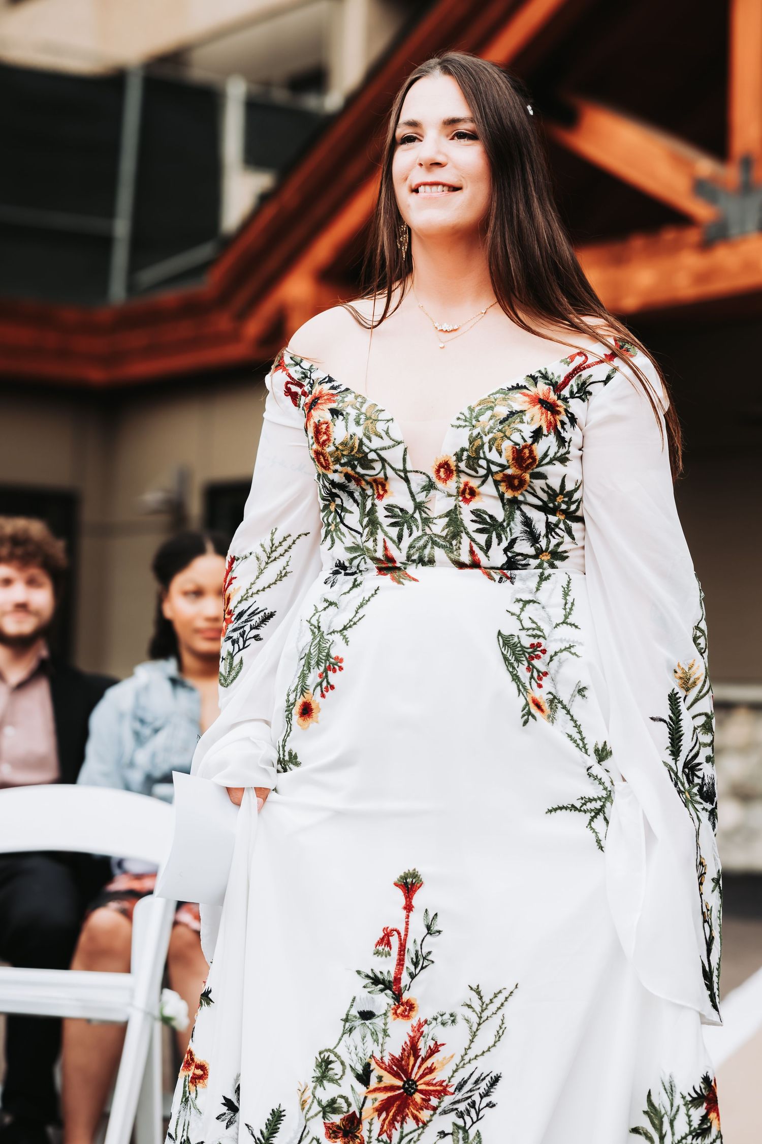 Young woman in white floral embroidered off-shoulder gown smiling at outdoor wedding ceremony.