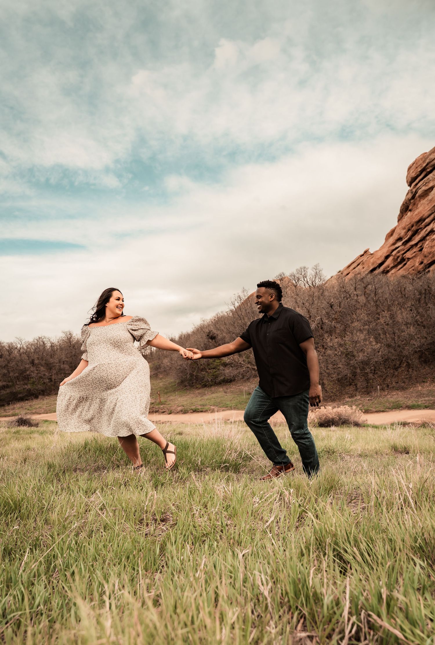 A joyful couple holds hands while dancing in a field near red rock formations.